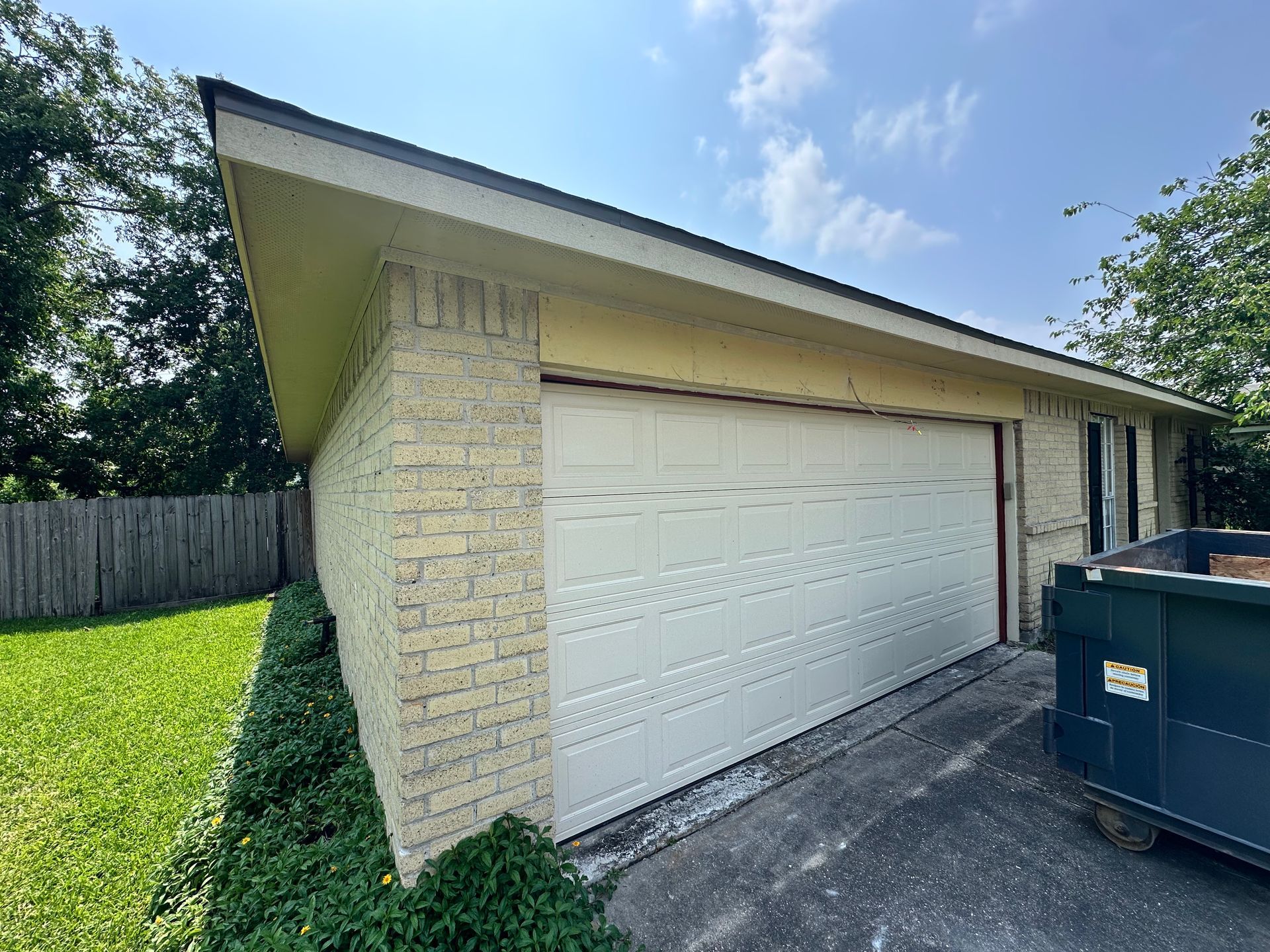Garage with beige brick exterior, white door, and a dumpster on a driveway, with a grassy lawn and a blue sky.