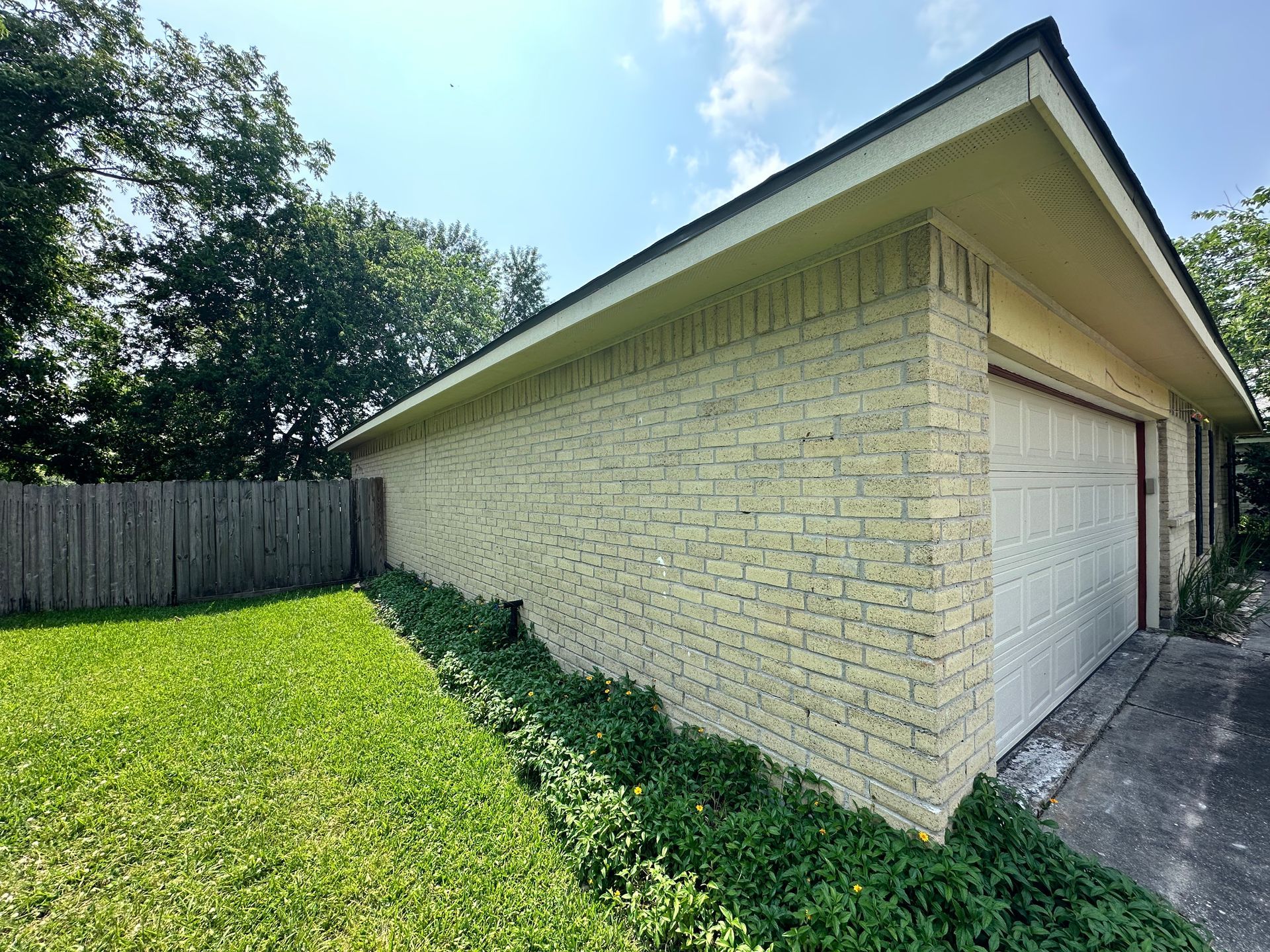 Tan brick garage with white door, next to a green lawn and wooden fence under a blue sky.