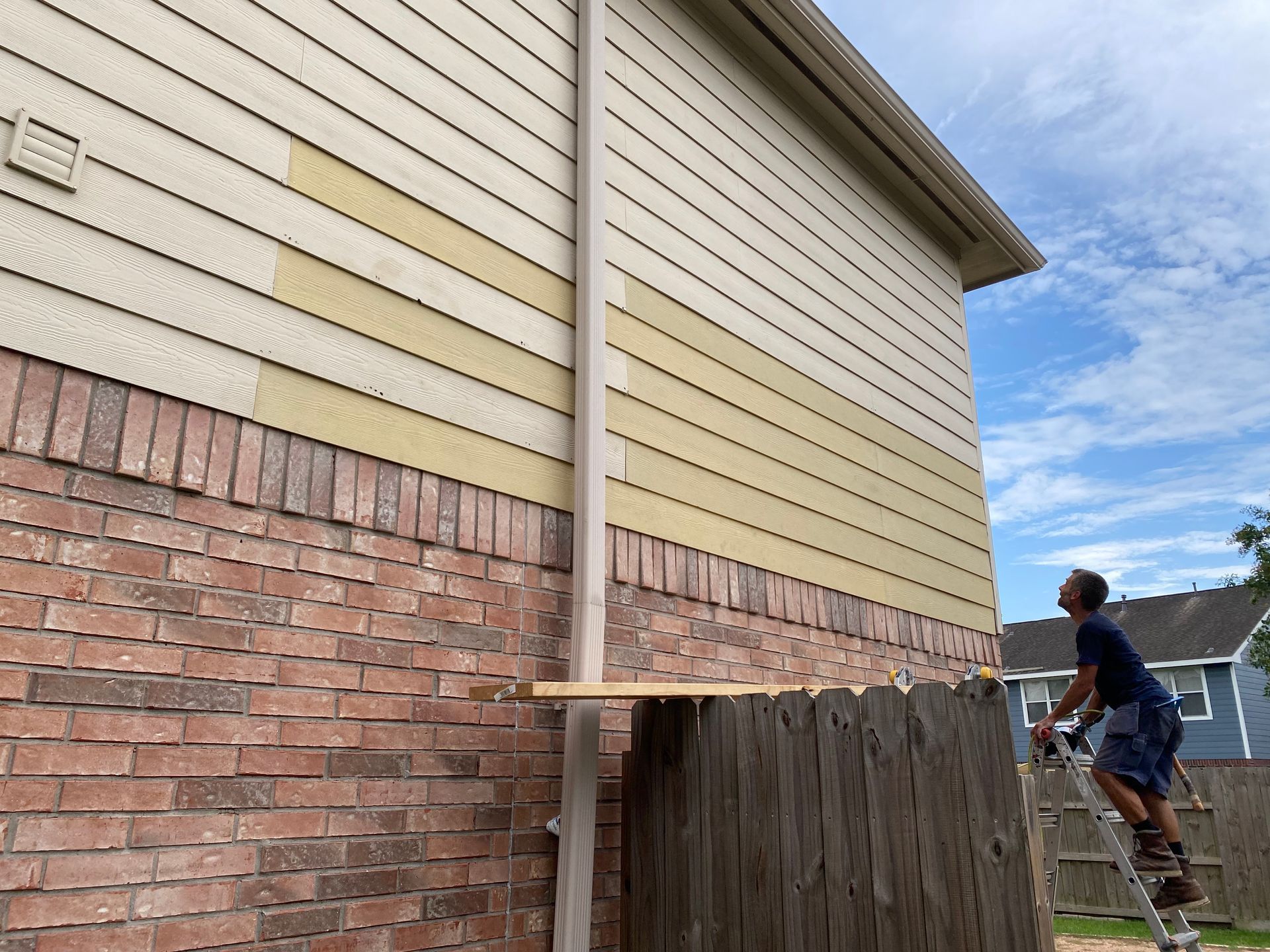 Person on ladder working on siding of a brick and wood house; blue sky.