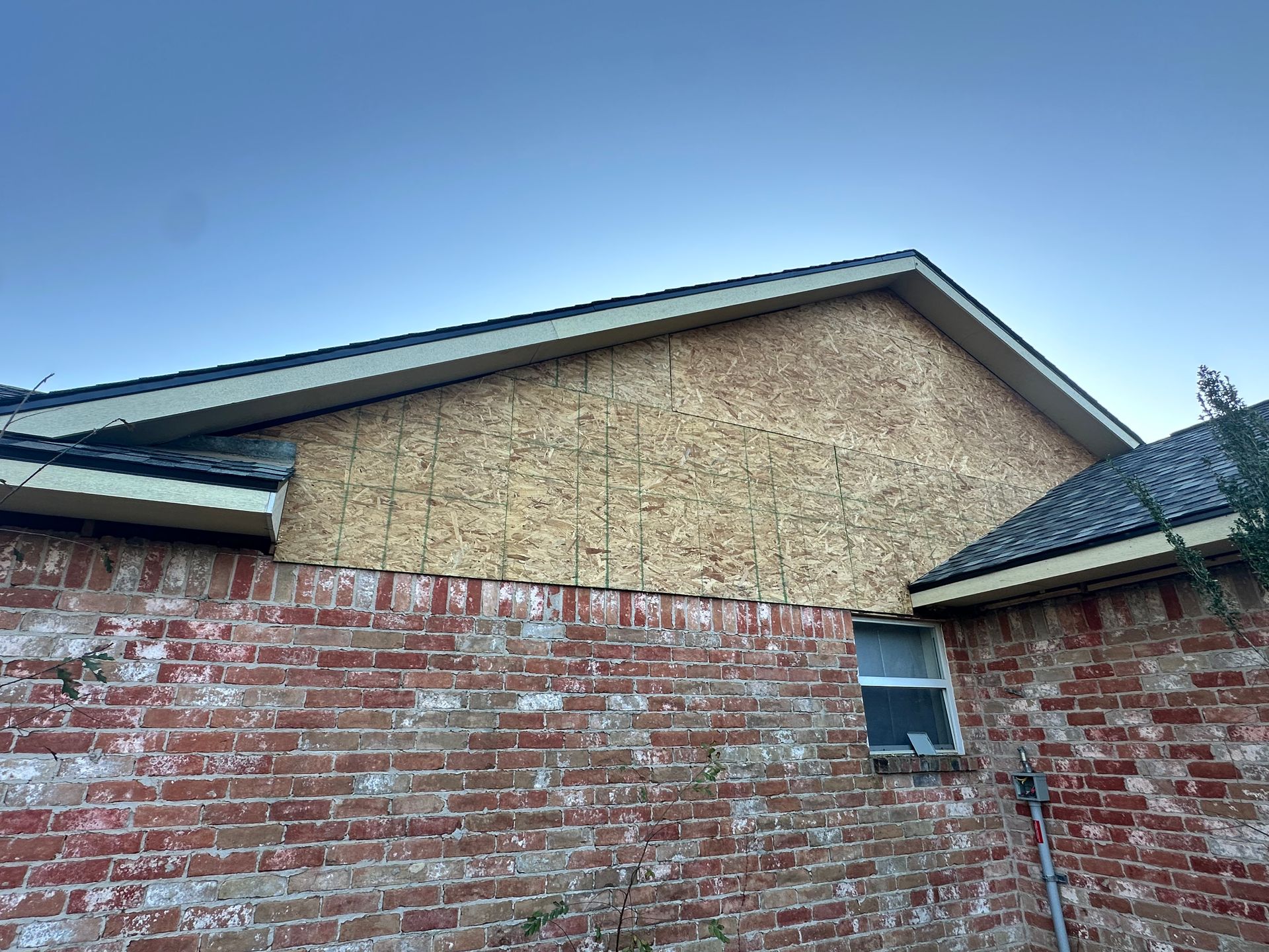 Brick wall with a section of OSB sheathing on a roof's gable end. Dark roof with beige trim against a blue sky.