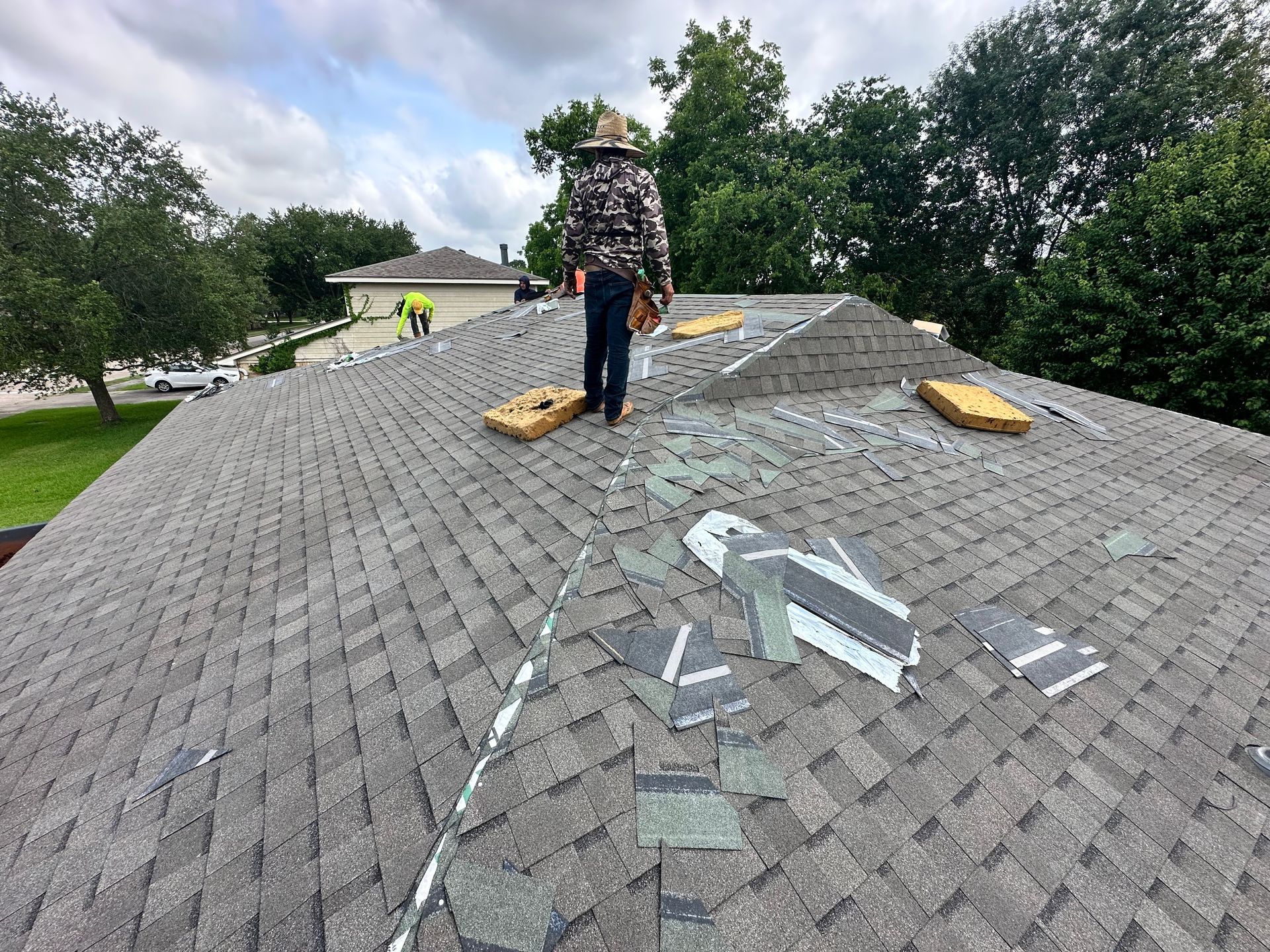 Roofer on a gray shingled roof, walking near removed shingles. Tools and trees visible in a residential setting.