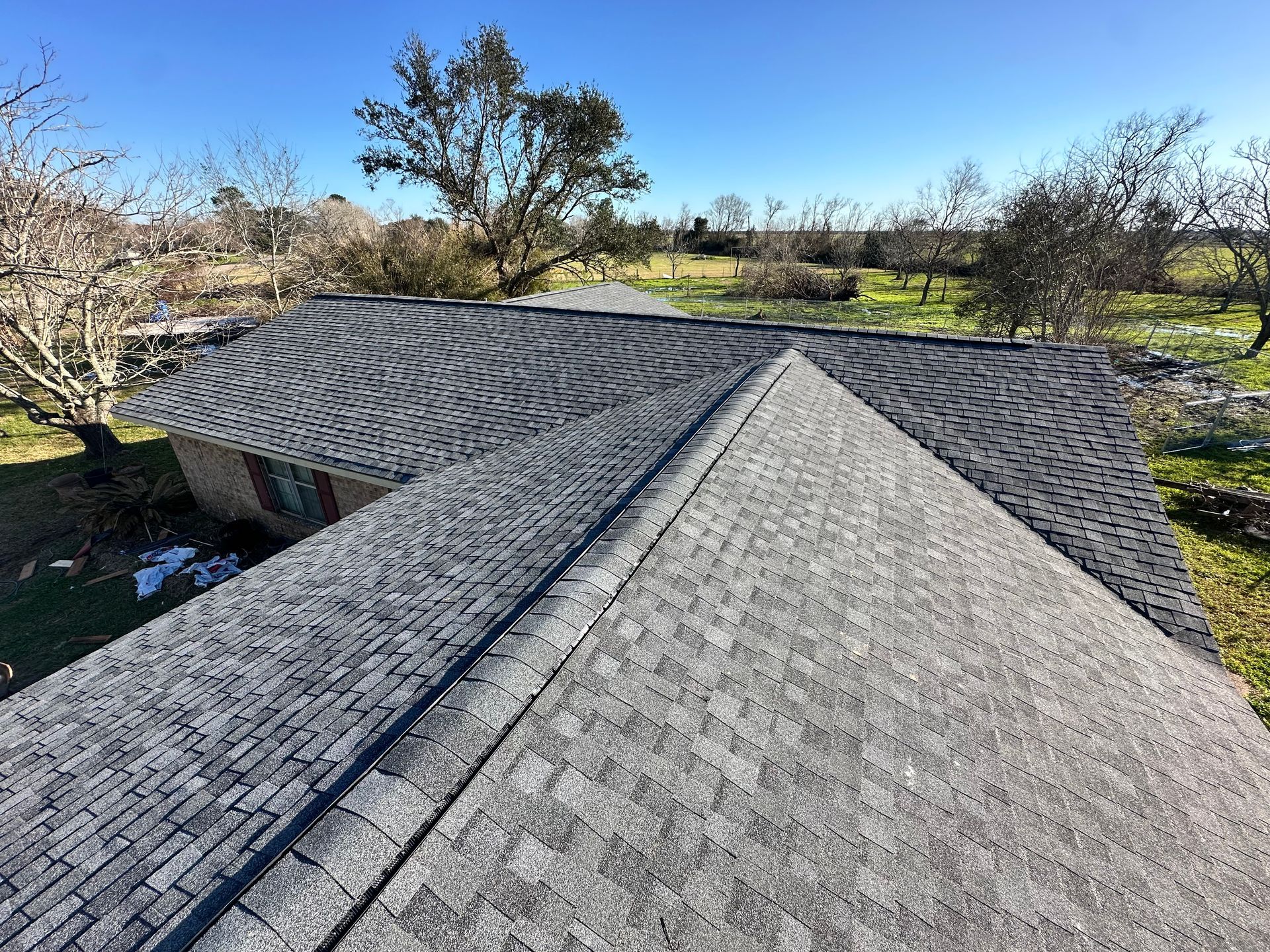 Overhead view of a house roof with dark gray shingles, trees, and blue sky.