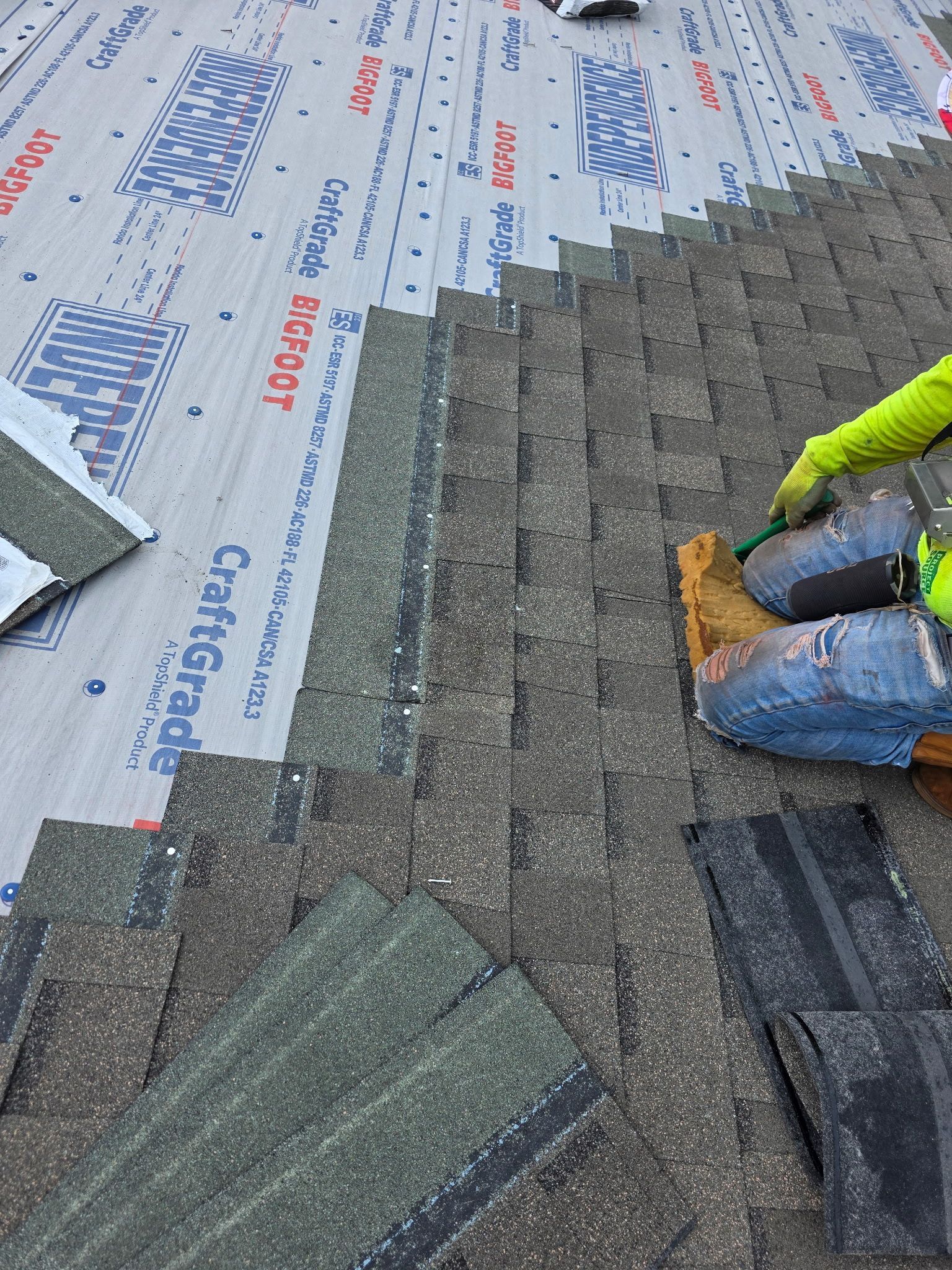 Roofer installing shingles on a roof. Green gloves, safety gear. Gray and brown shingles, blue underlayment.