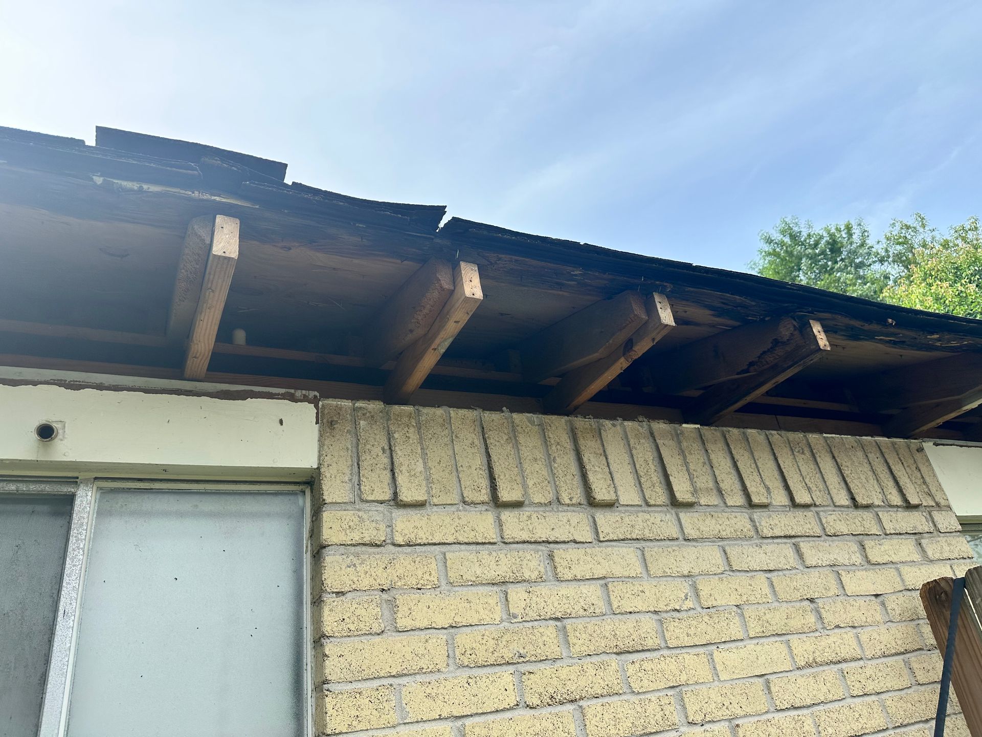 Damaged roof overhang above a brick wall and window, with exposed wood and missing shingles.