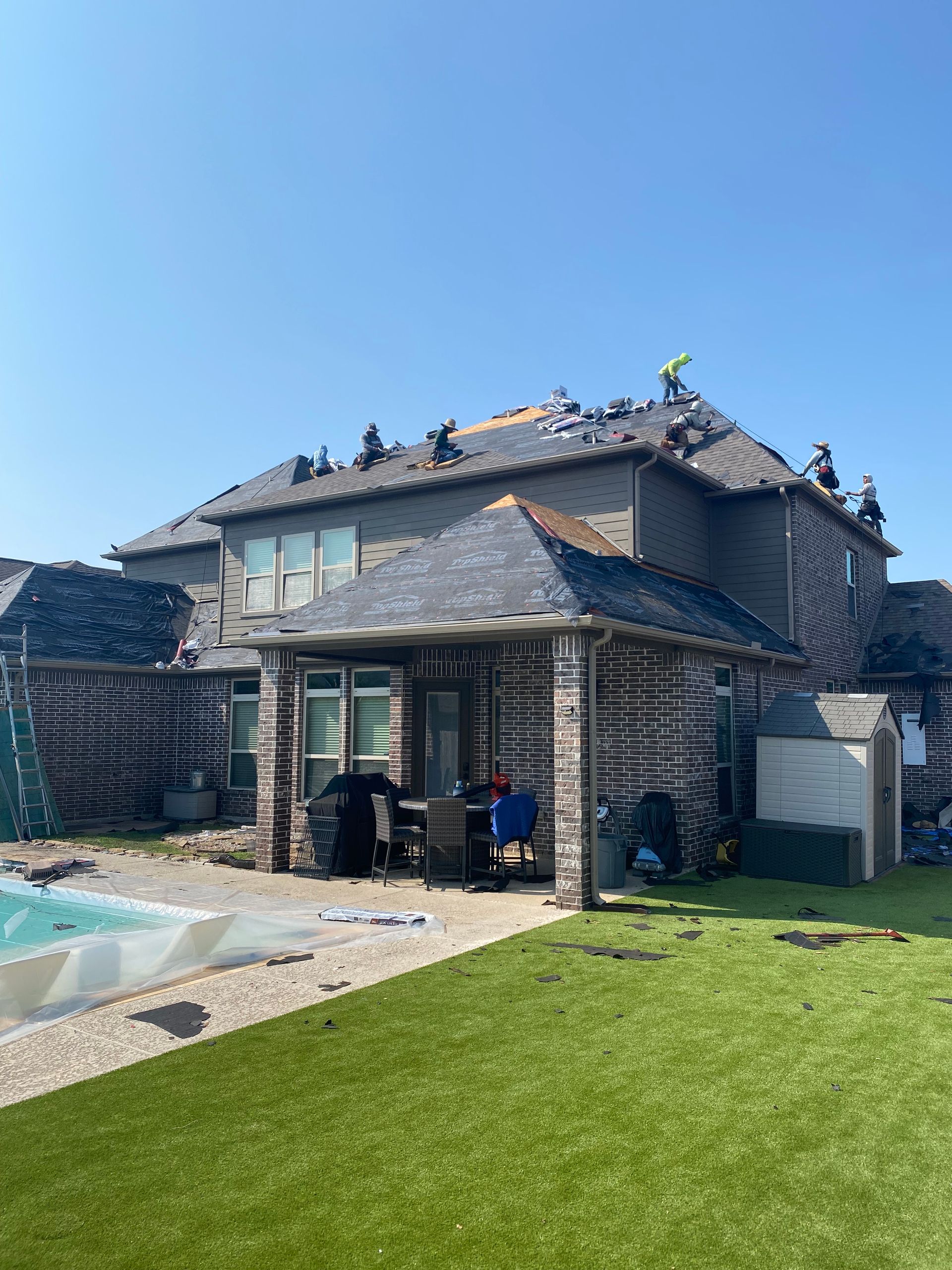 Roofers working on a residential roof under a clear, blue sky. Backyard with pool and lawn.