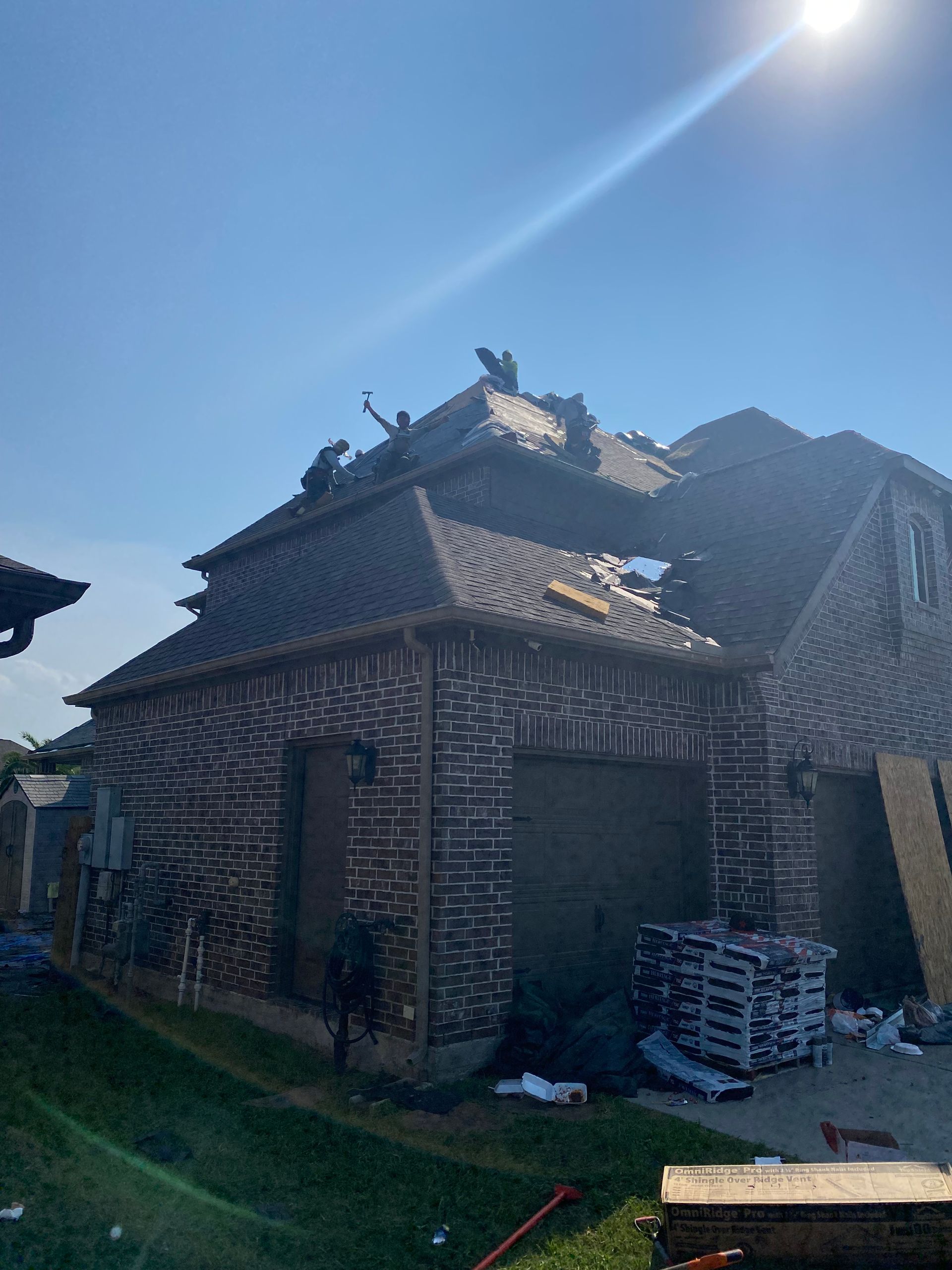 House with roof damage, likely under repair; dark brick exterior, clear sunny sky.