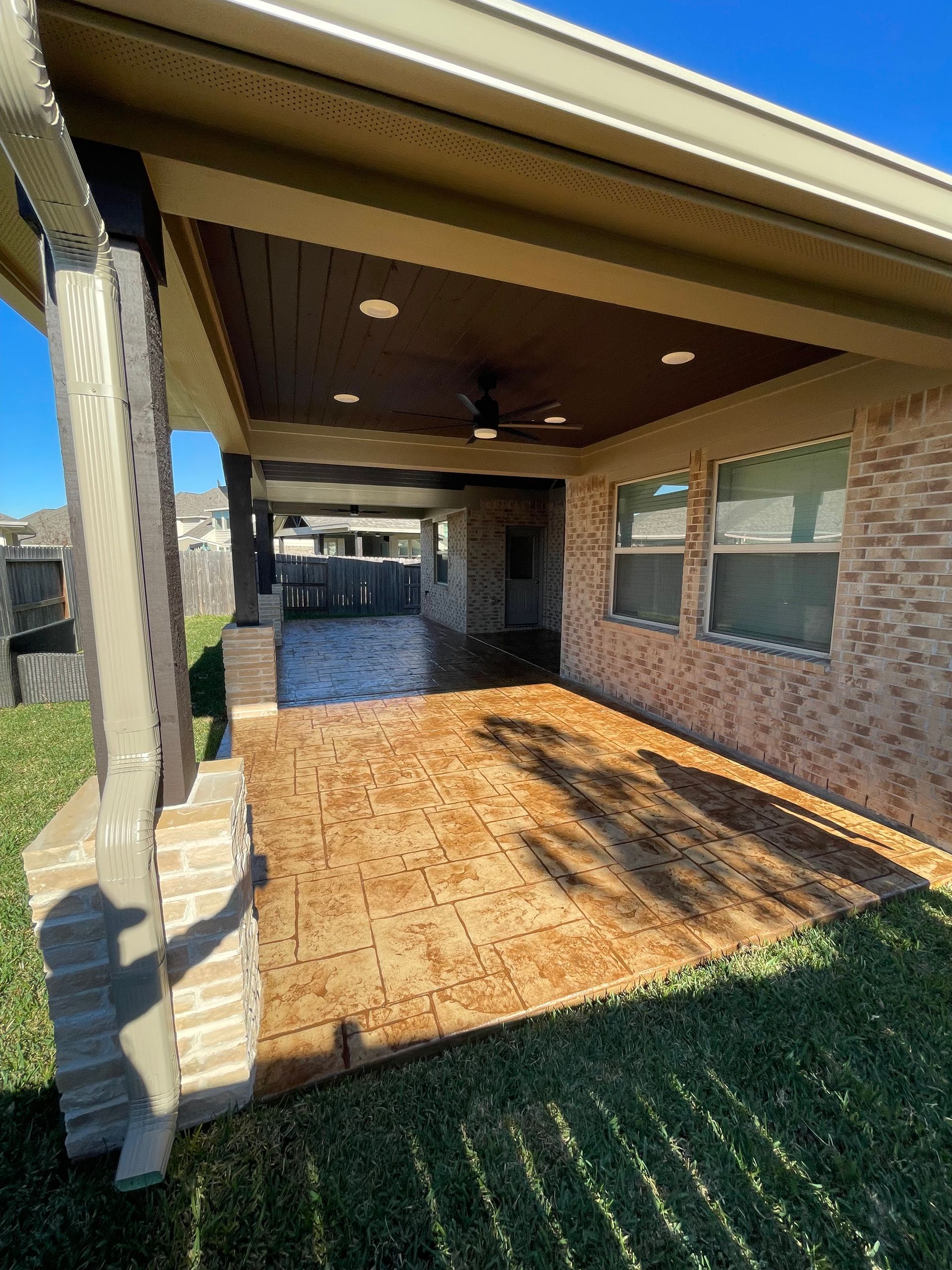 Covered patio with stamped concrete floor, brick wall, and dark ceiling, with grass and blue sky.