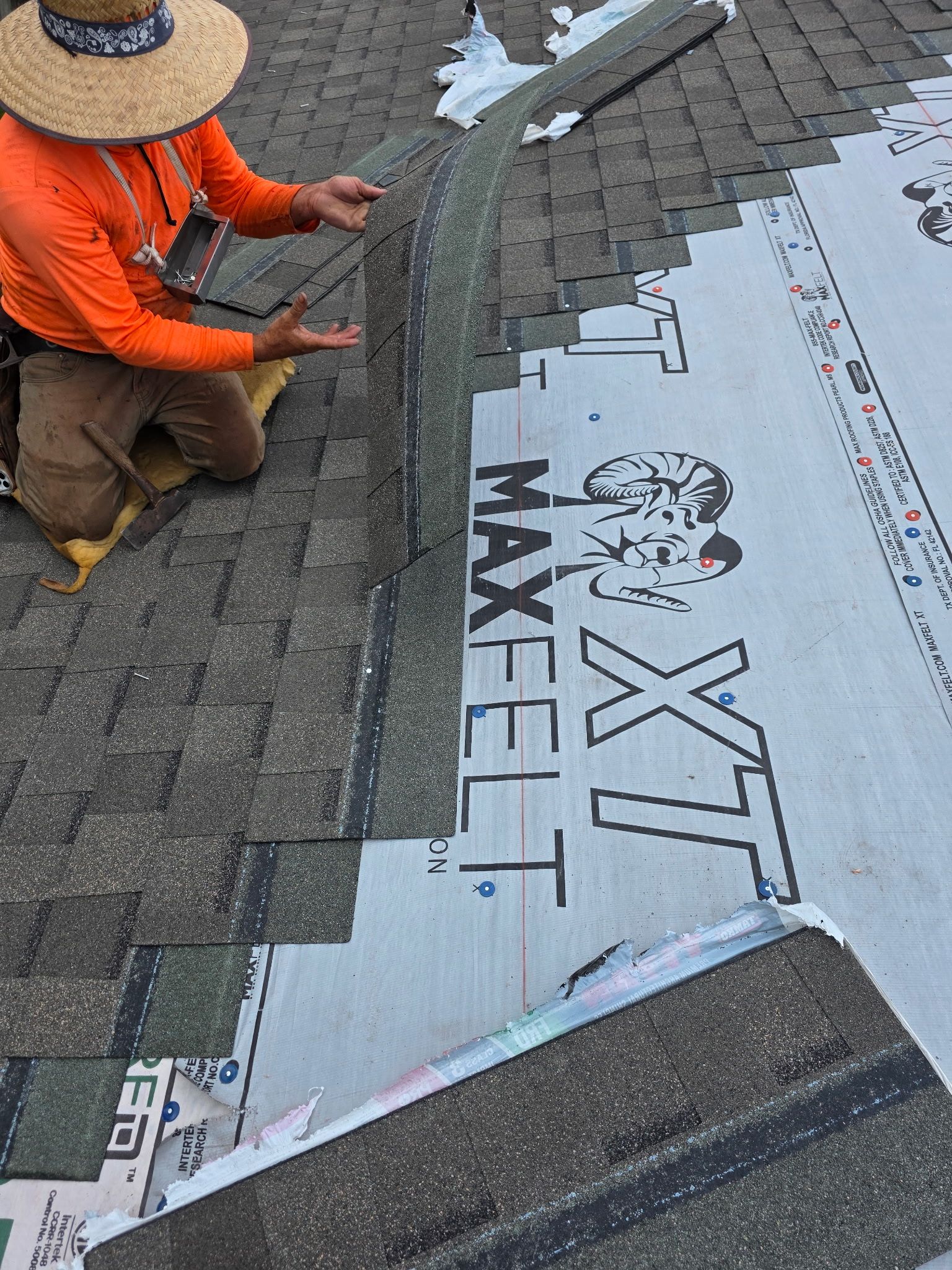Roofer kneeling on a roof, installing shingles. They wear a straw hat and orange shirt, pointing at a shingle edge.