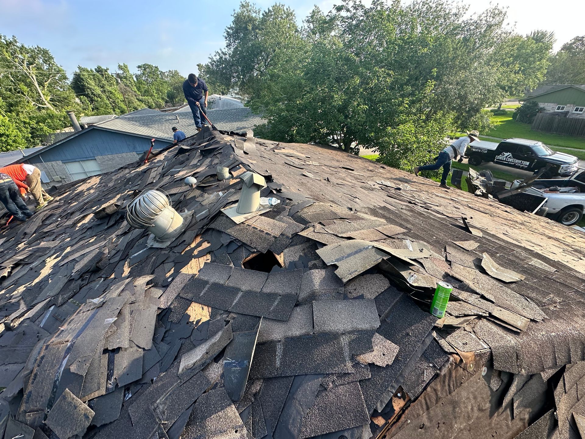 Workers removing shingles from a roof. Several people are on the roof, debris is scattered, and trees and cars are in the background.