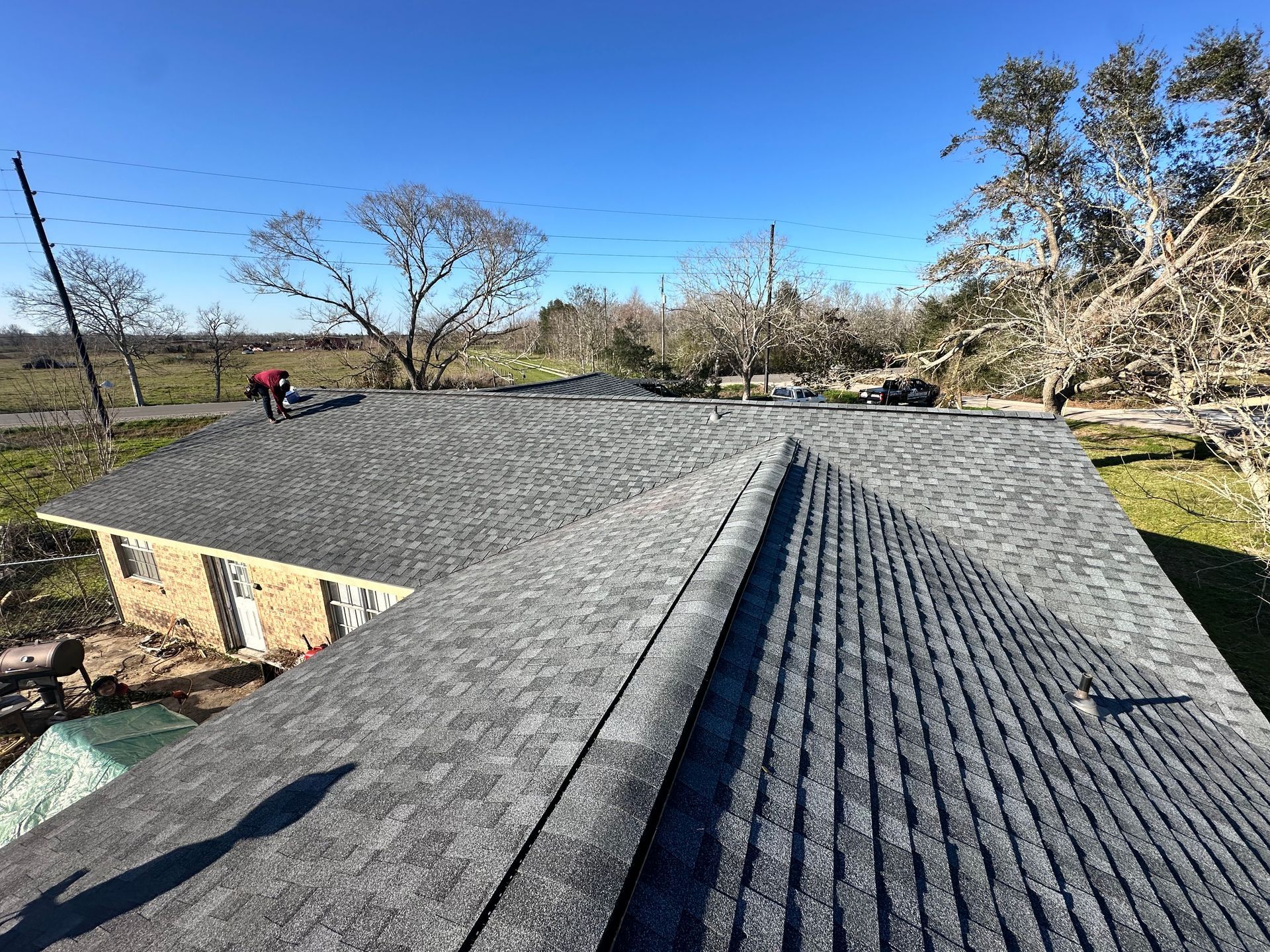 A person on a dark gray shingle roof under a clear blue sky. Trees and a building are in the background.
