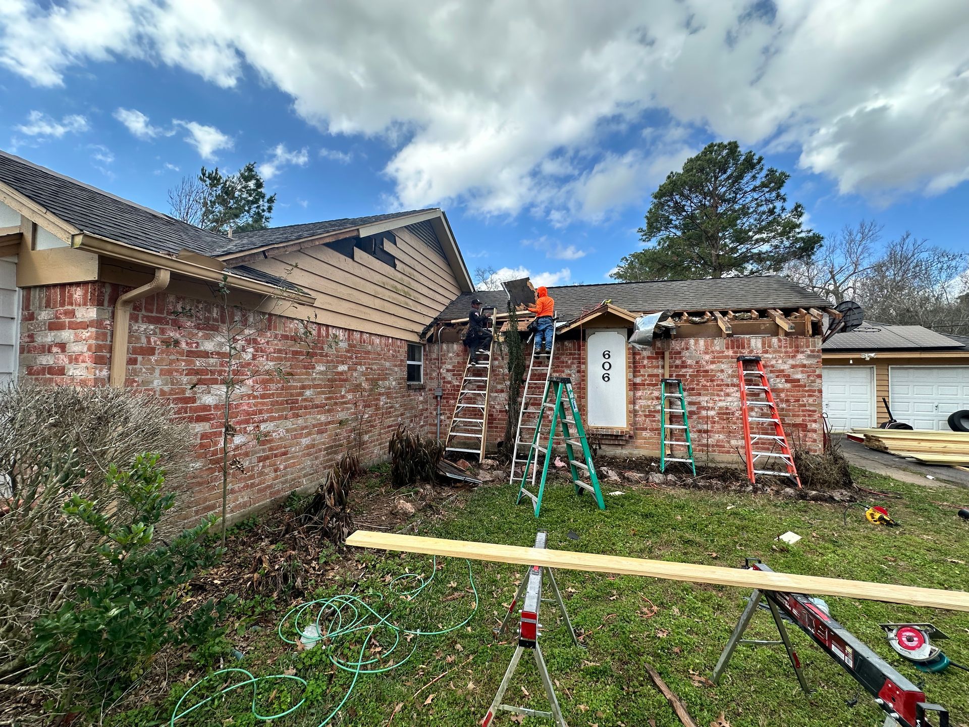 House under construction: workers on ladders, removing roofing and siding from brick building. Sunny day.