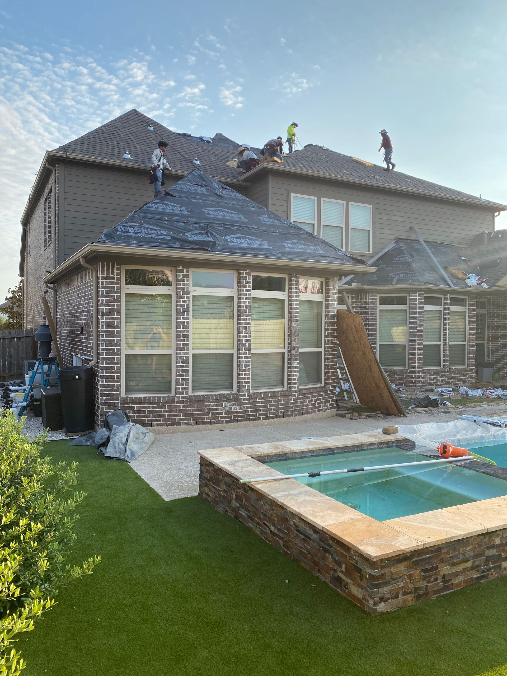 Roofers working on a home's roof, near a swimming pool. Brick exterior, blue sky.