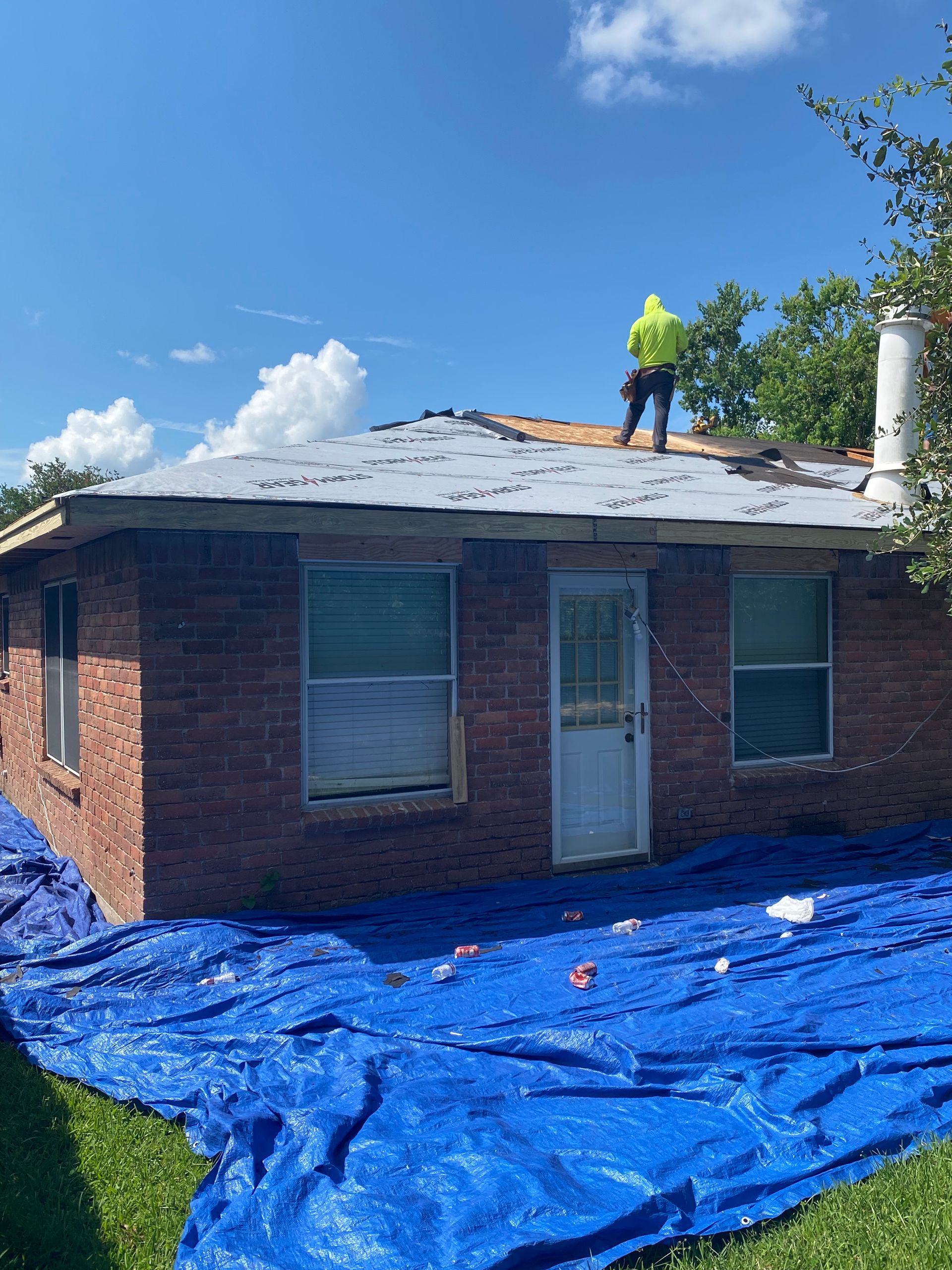 Person on a roof replacing shingles, brick building, blue tarp on ground, sunny day.