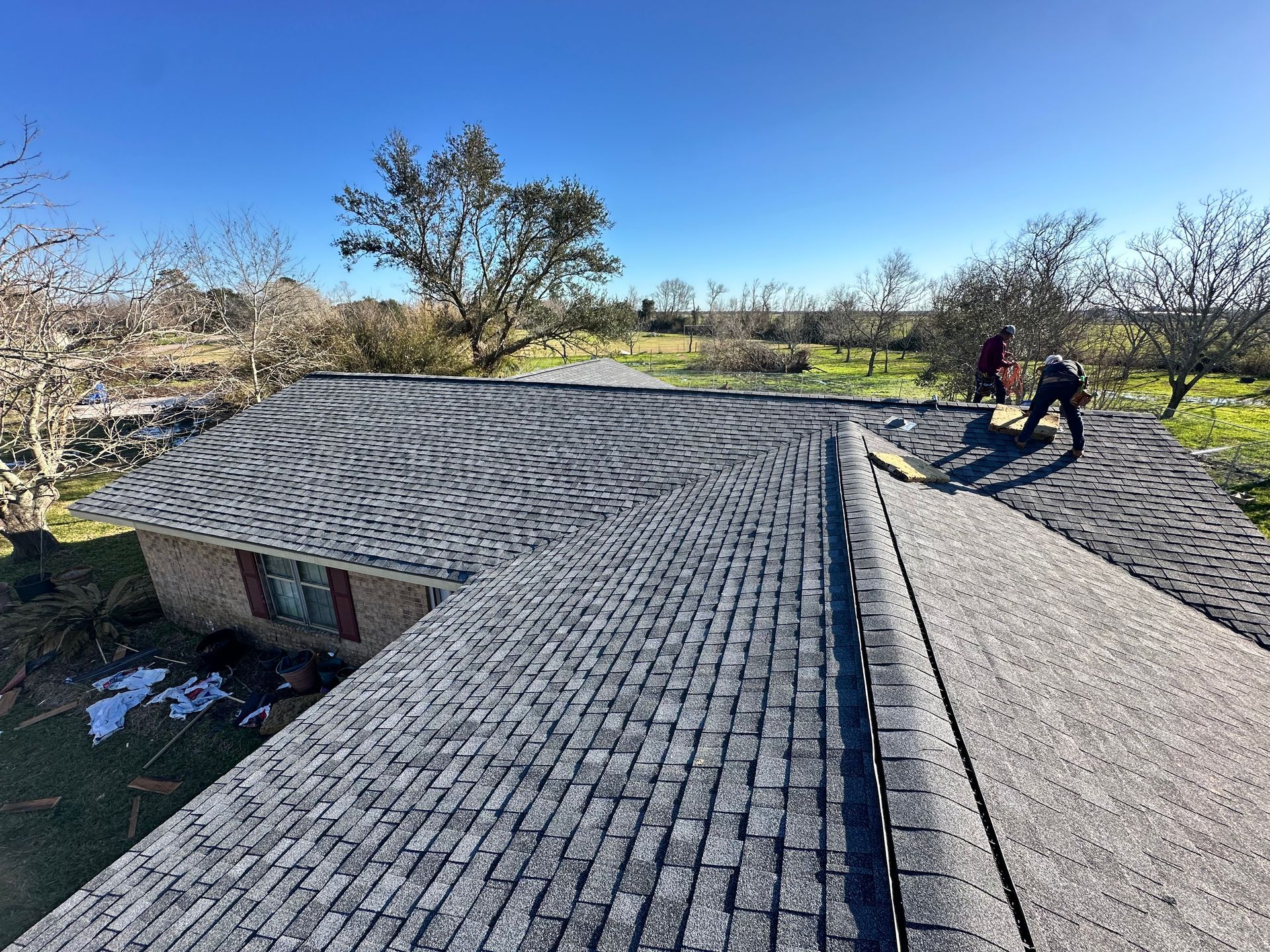 Workers on a dark shingled roof under a clear blue sky, with a house and trees in the background.