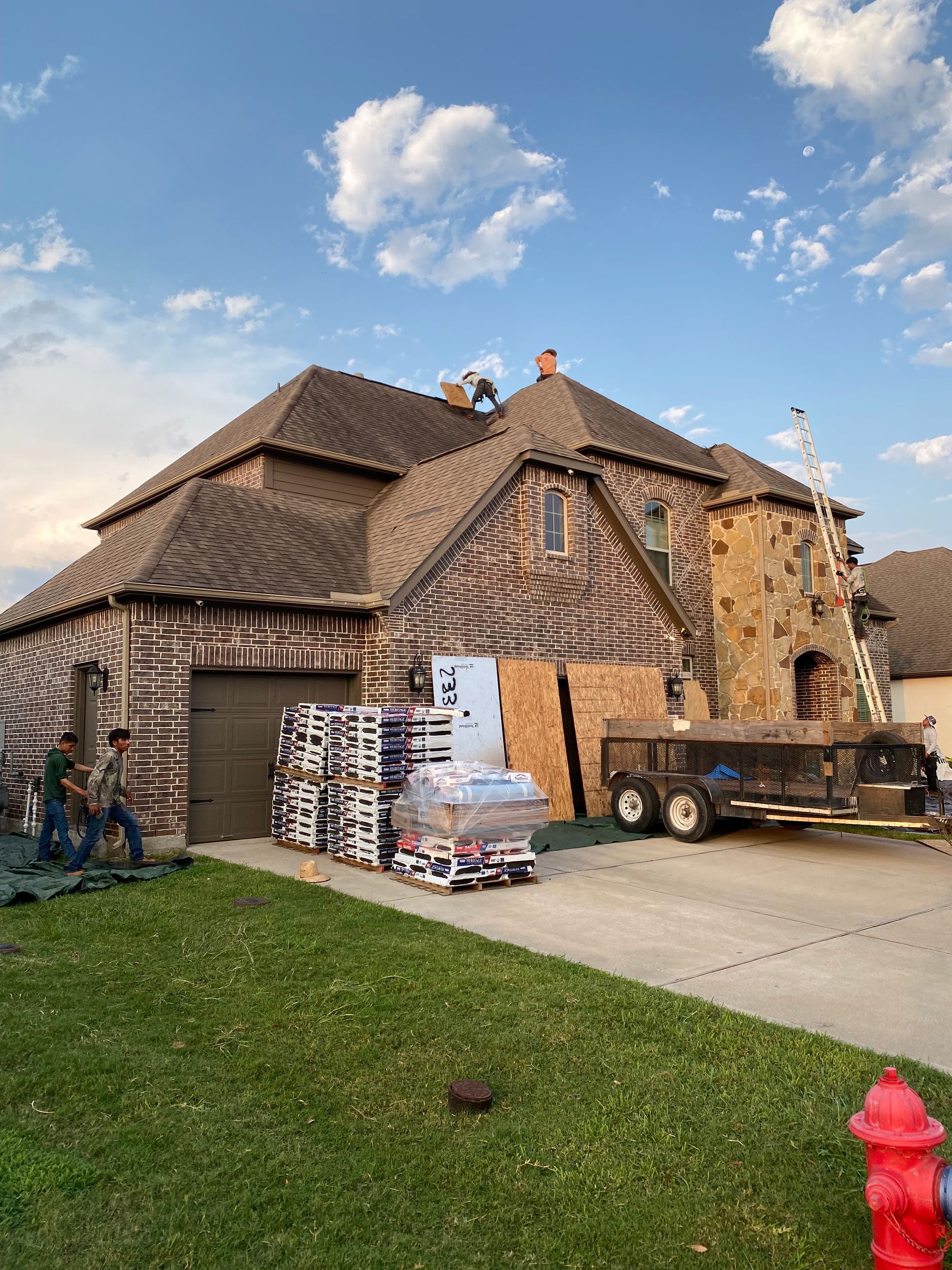 House with roofing materials, trailer, and ladder. Green lawn and blue sky.