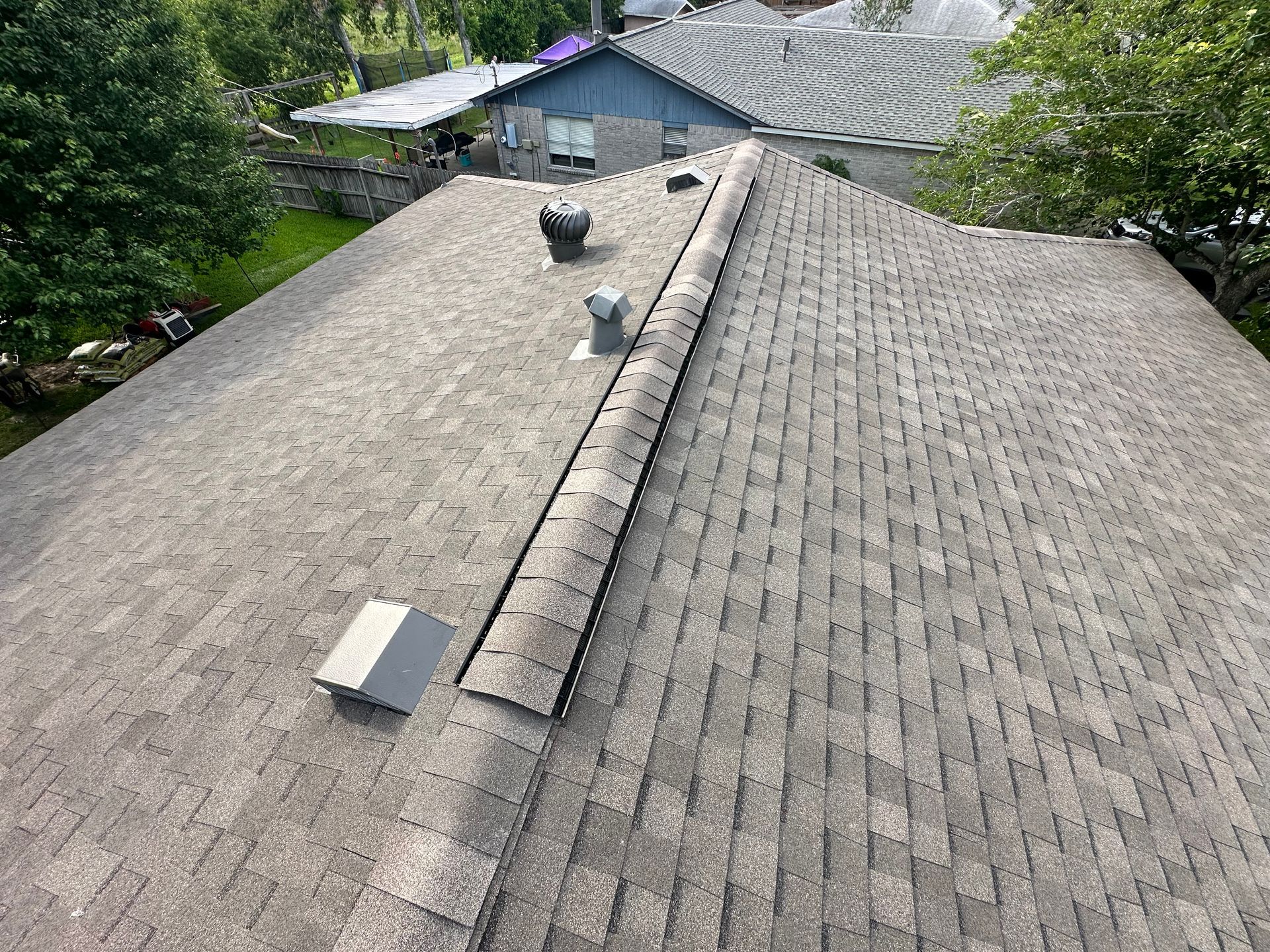 Overhead view of a gray shingle roof with vents and a ridge cap, surrounded by trees and buildings.