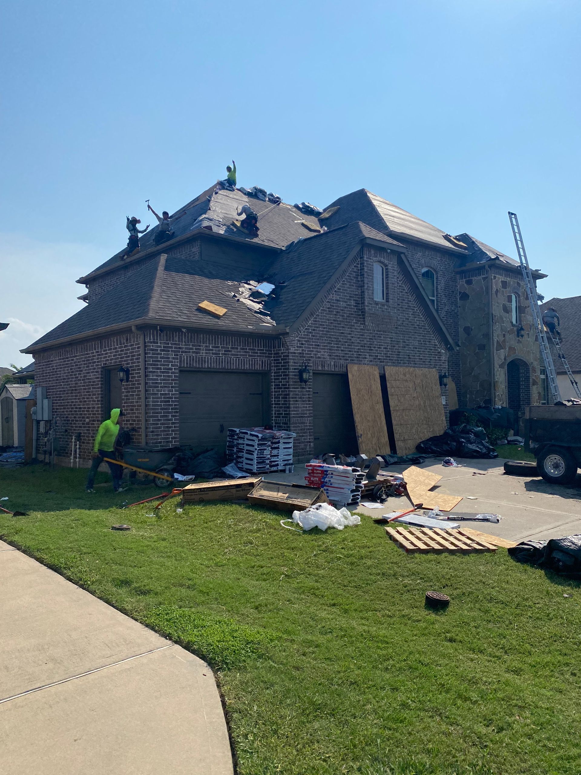 House with roof damage being repaired; workers on roof. Debris and materials on ground. Sunny day.