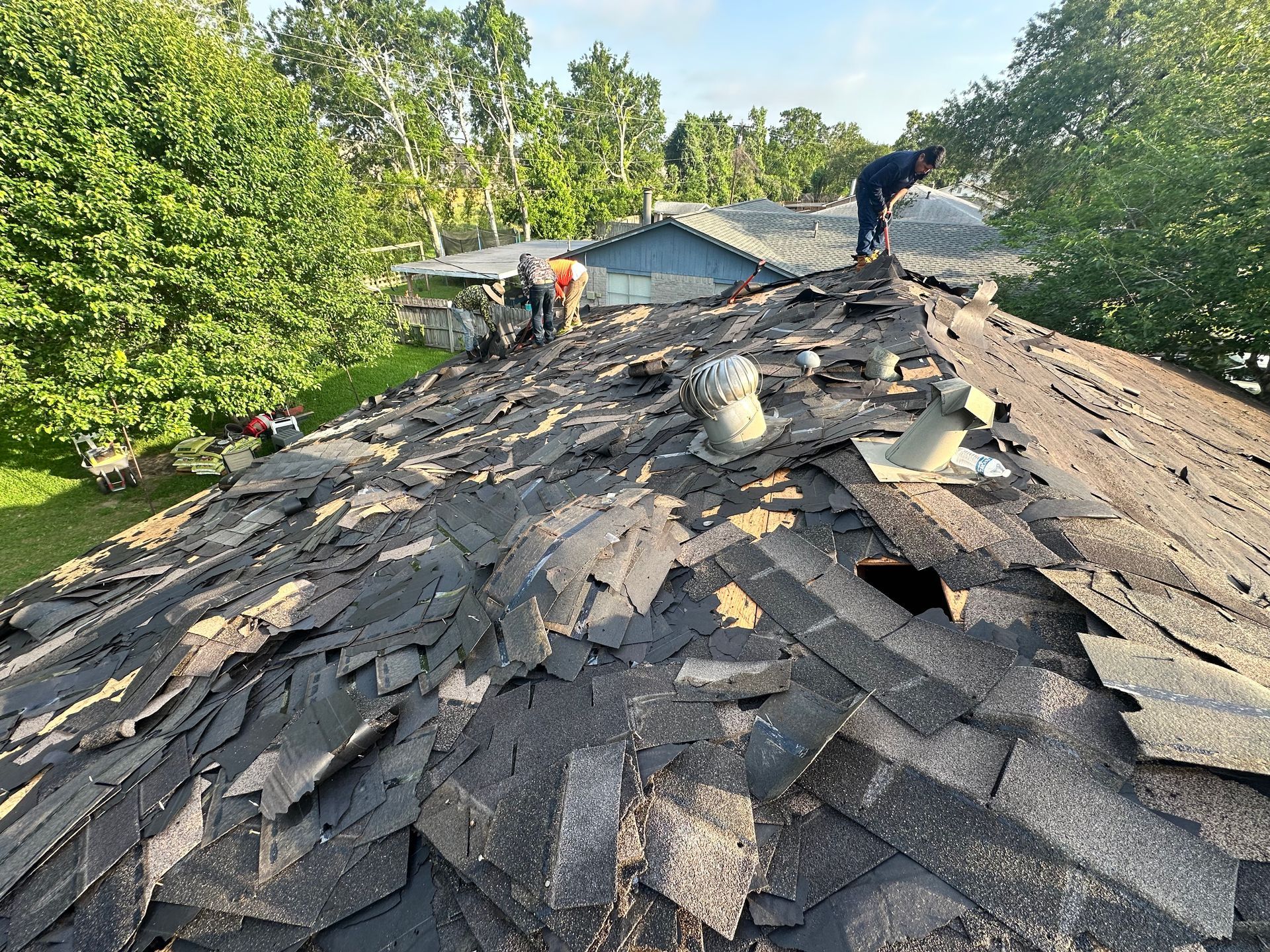 Roof being torn off with workers on top; trees and houses in the background.