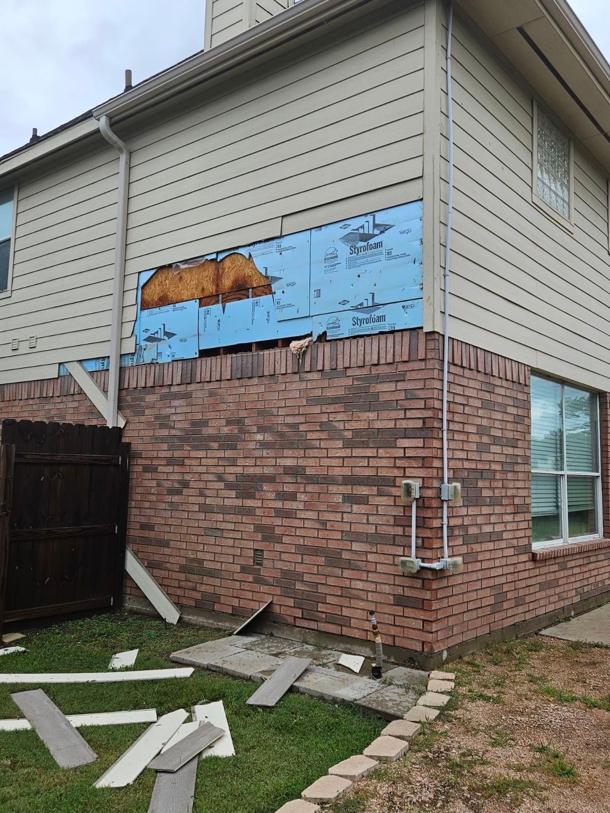 Exterior house corner with damaged siding above brick foundation; exposed blue sheathing.