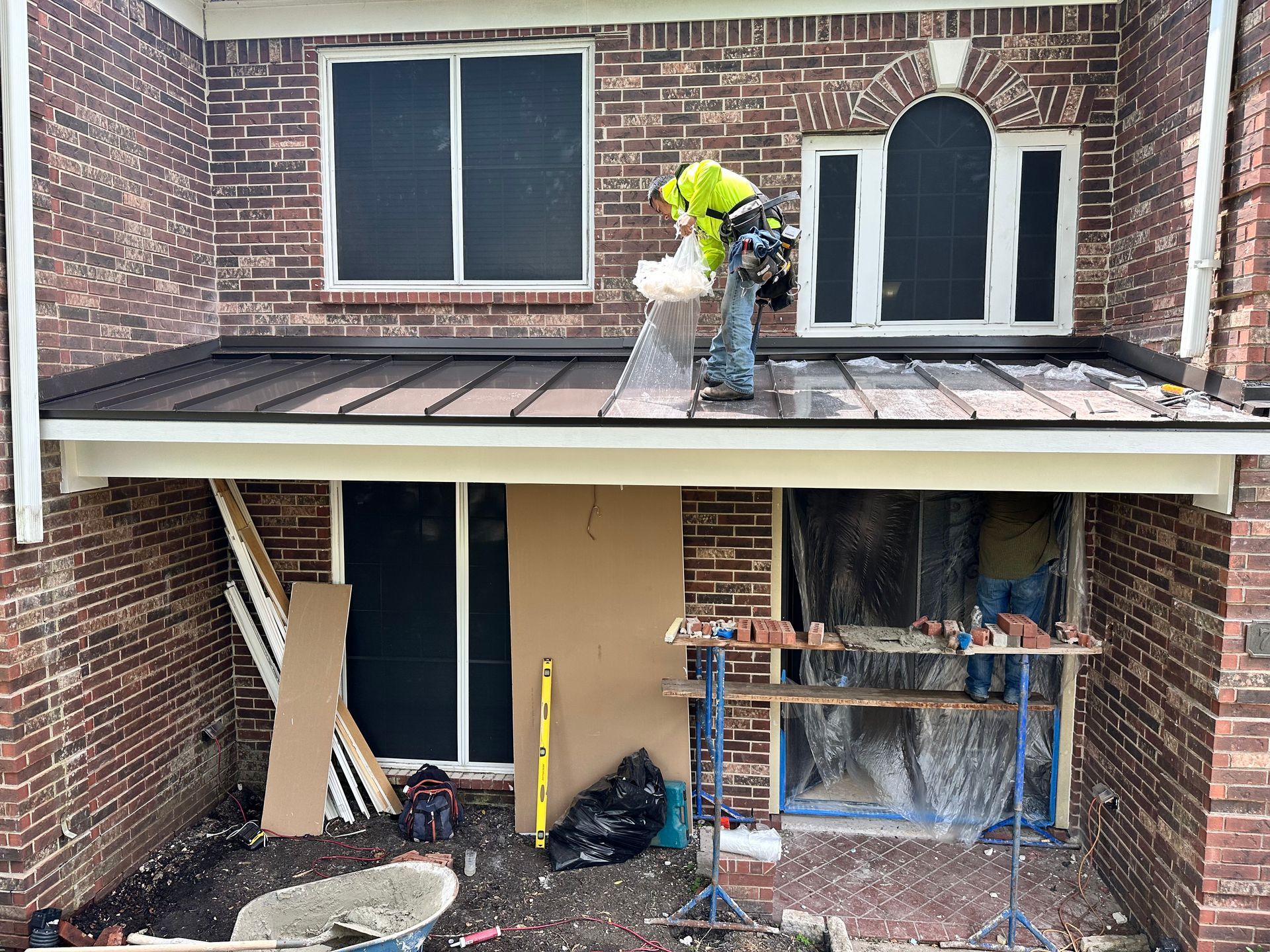 Construction site: worker on roof, brick building, windows covered, materials scattered below.