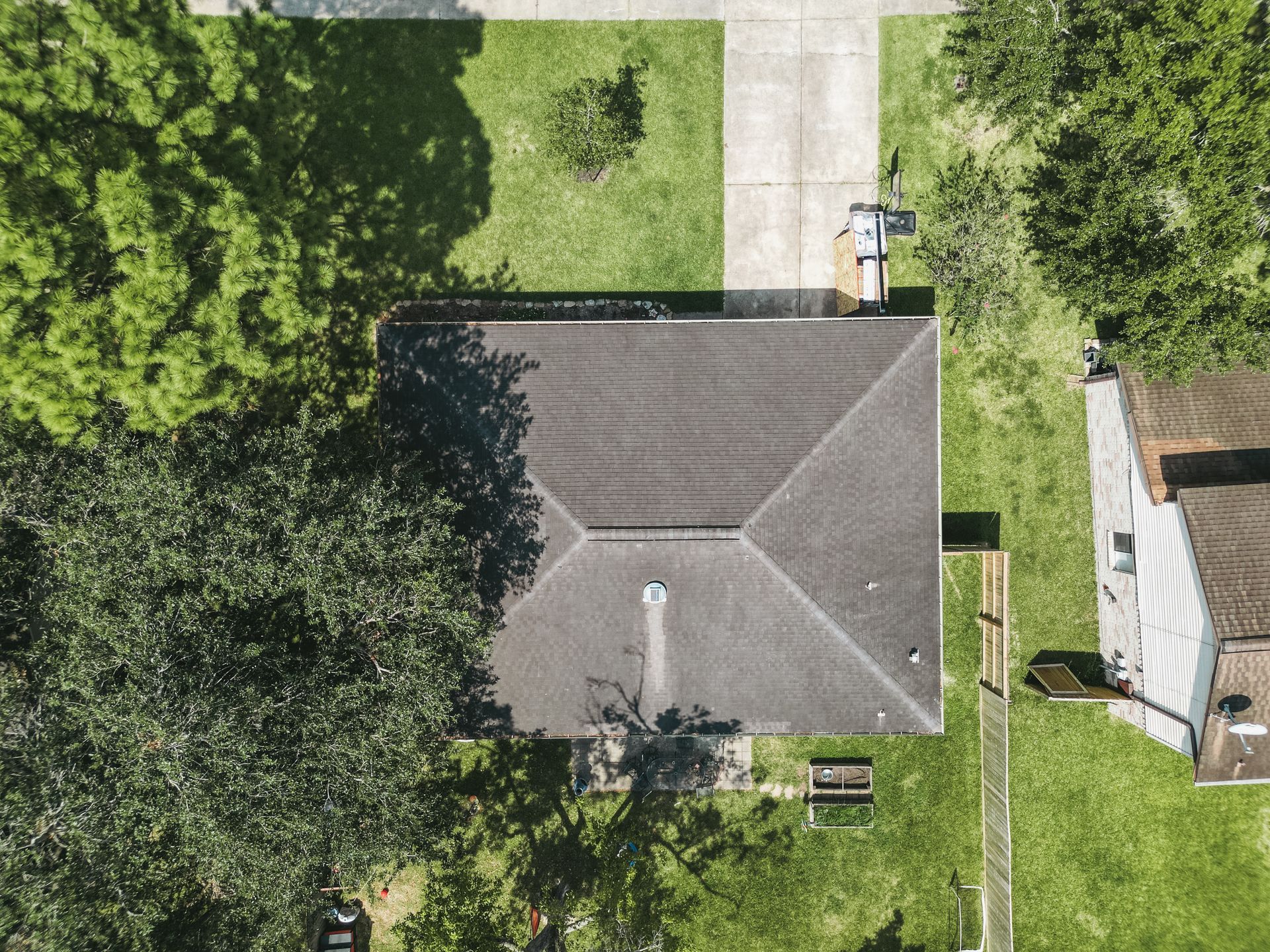Overhead view of a house with a gray roof, surrounded by green trees and grass. A concrete driveway is visible.