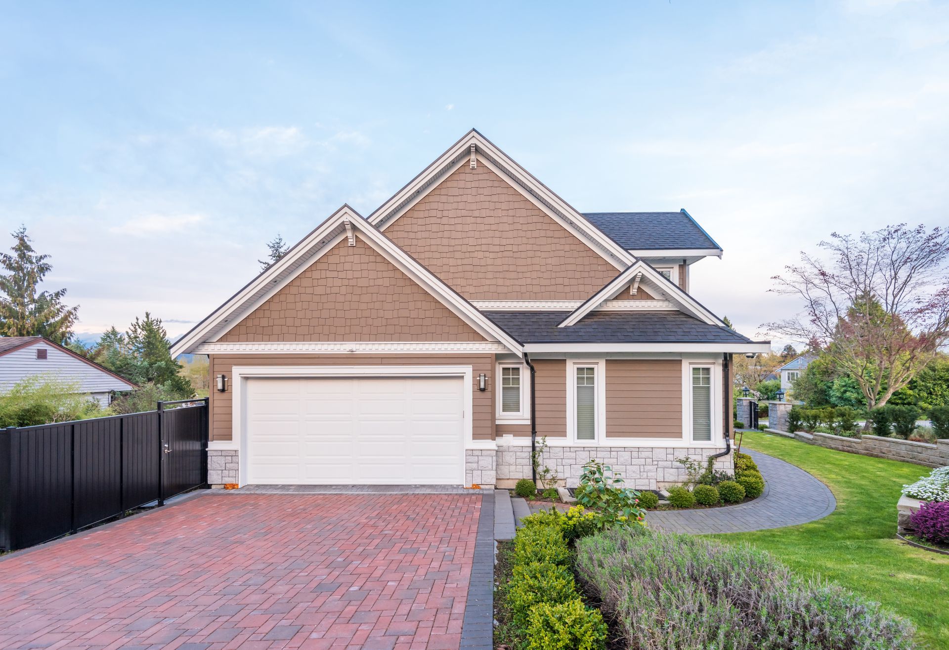 Tan and white house with brick driveway, garage door, and landscaping under a blue sky.