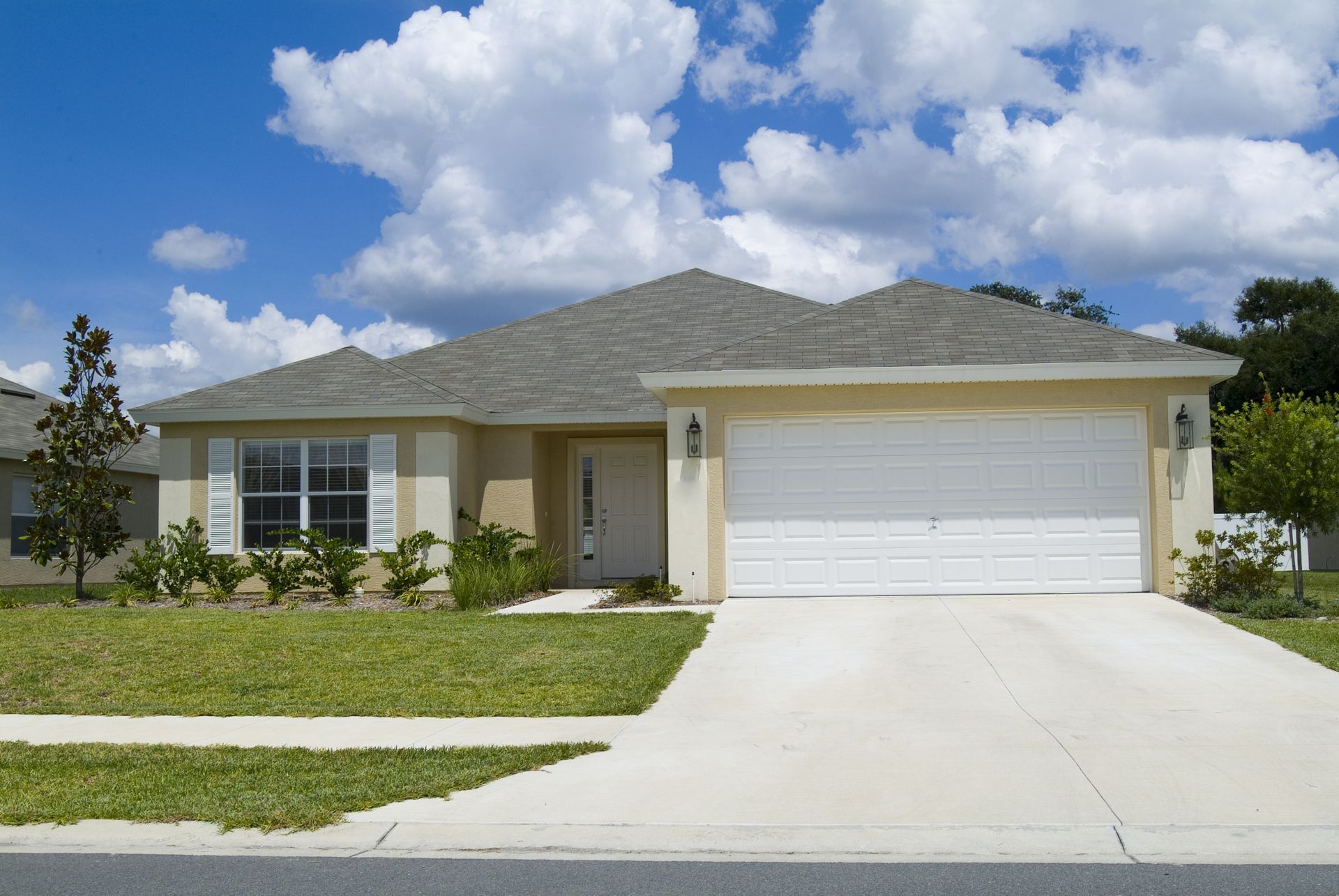 Tan one-story house with a two-car garage, white door, and green lawn under a blue sky with clouds.