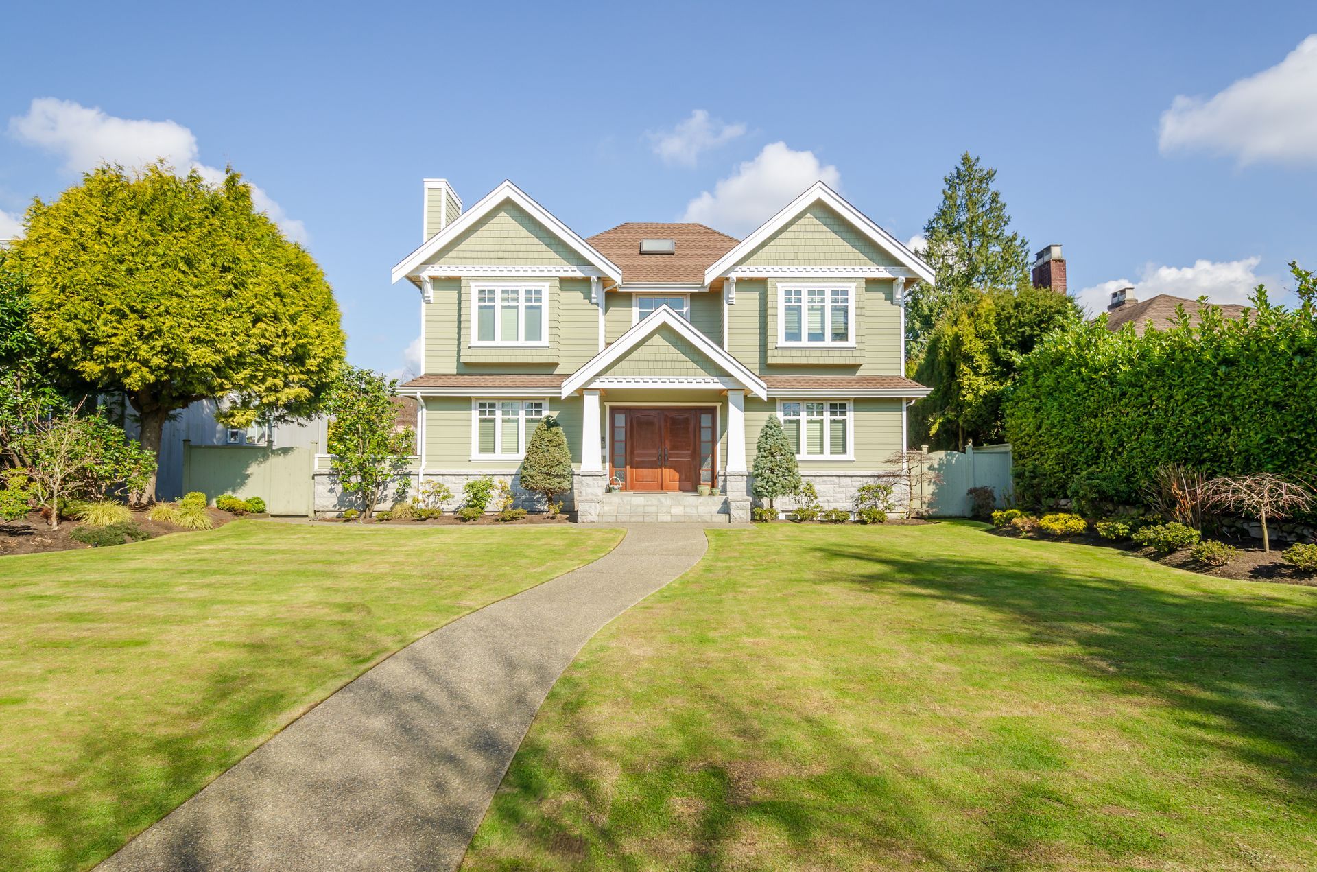 Green two-story house with brown door, surrounded by lawn and trees, under blue sky.