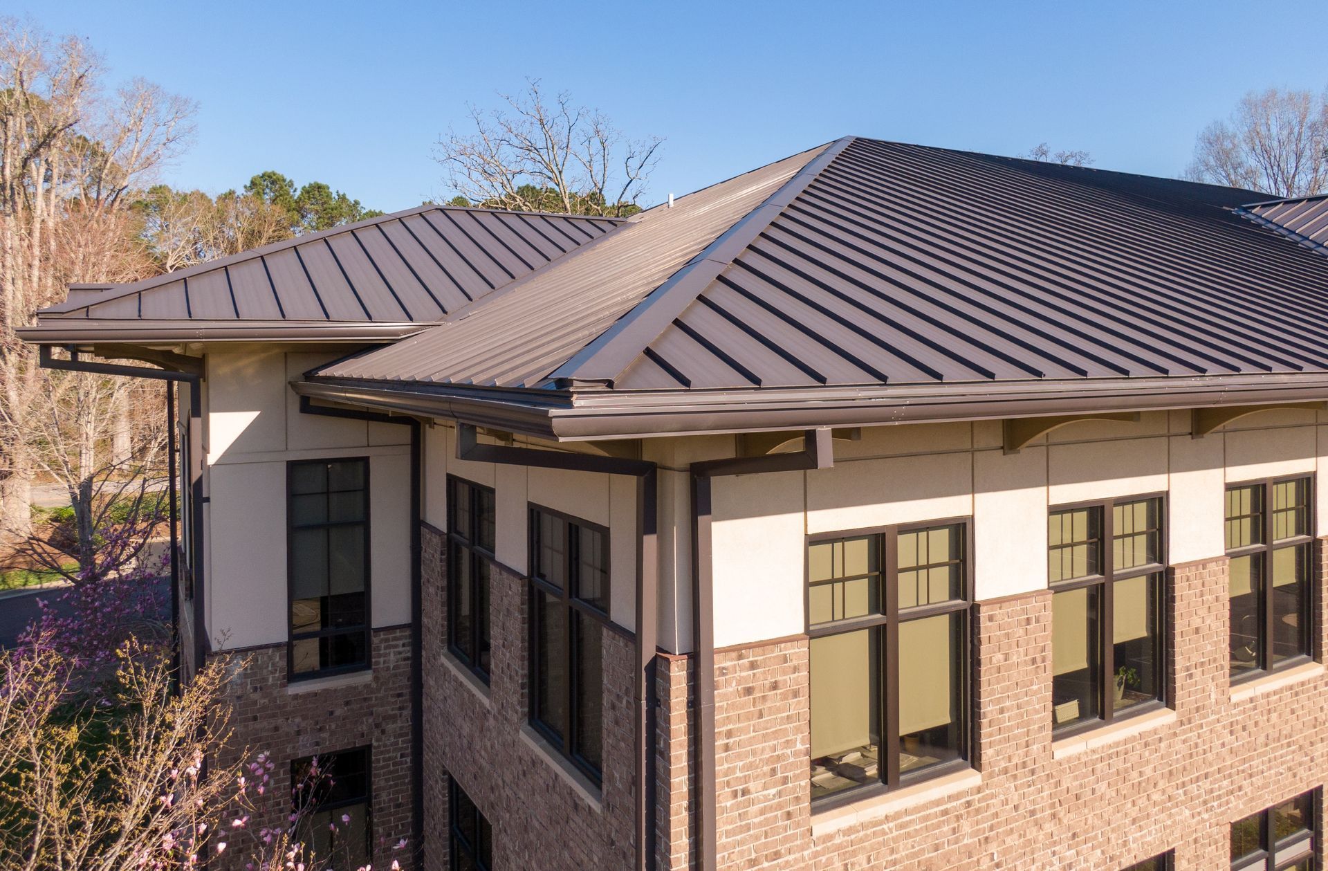 Brown metal roof on a light brick building with dark framed windows. Clear, sunny sky.