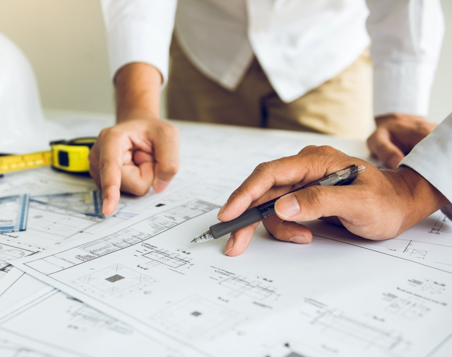 Hands pointing at an architectural blueprint on a desk with a pencil and tape measure nearby