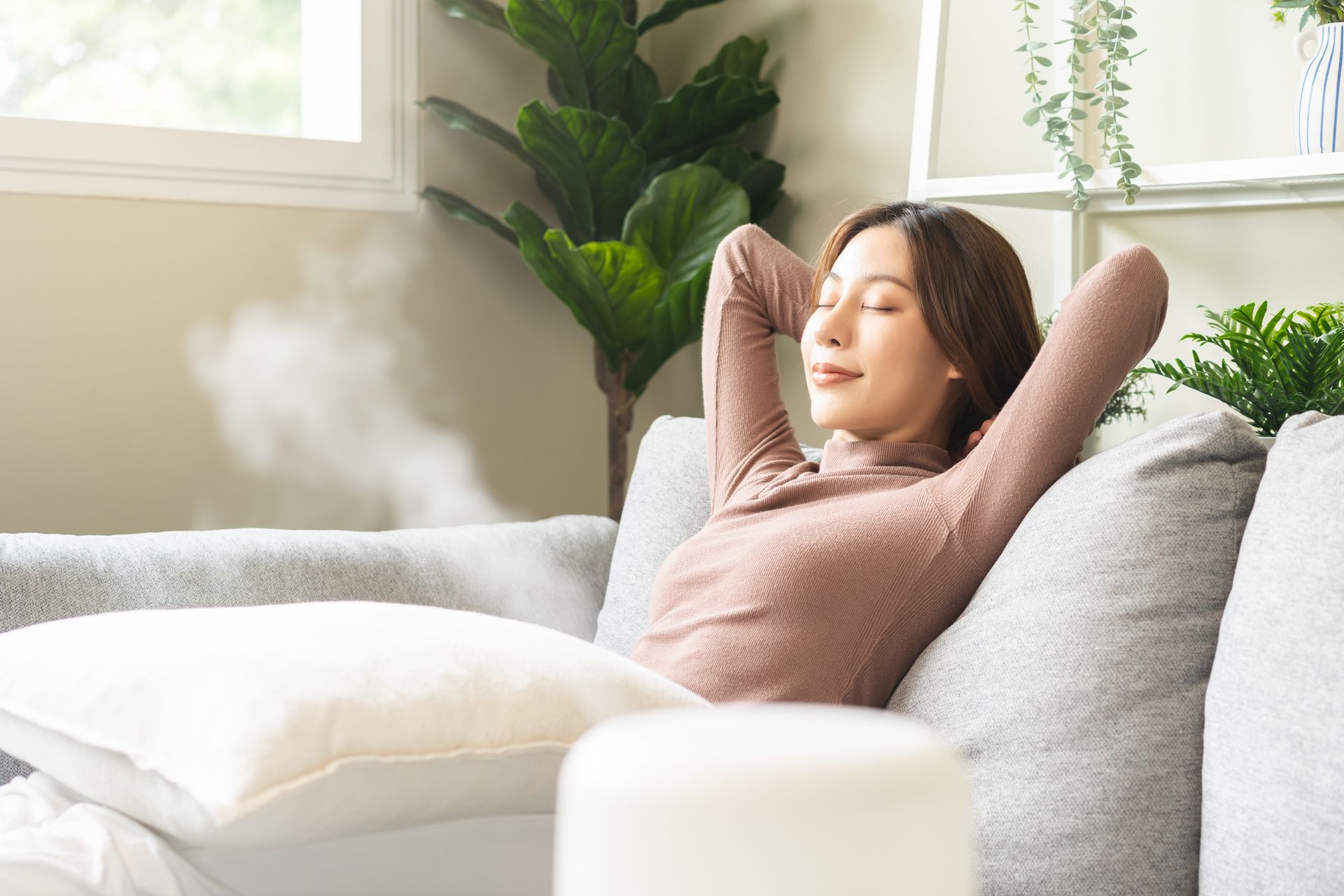 Woman relaxing on a couch, arms behind head, near a humidifier, with indoor plants.
