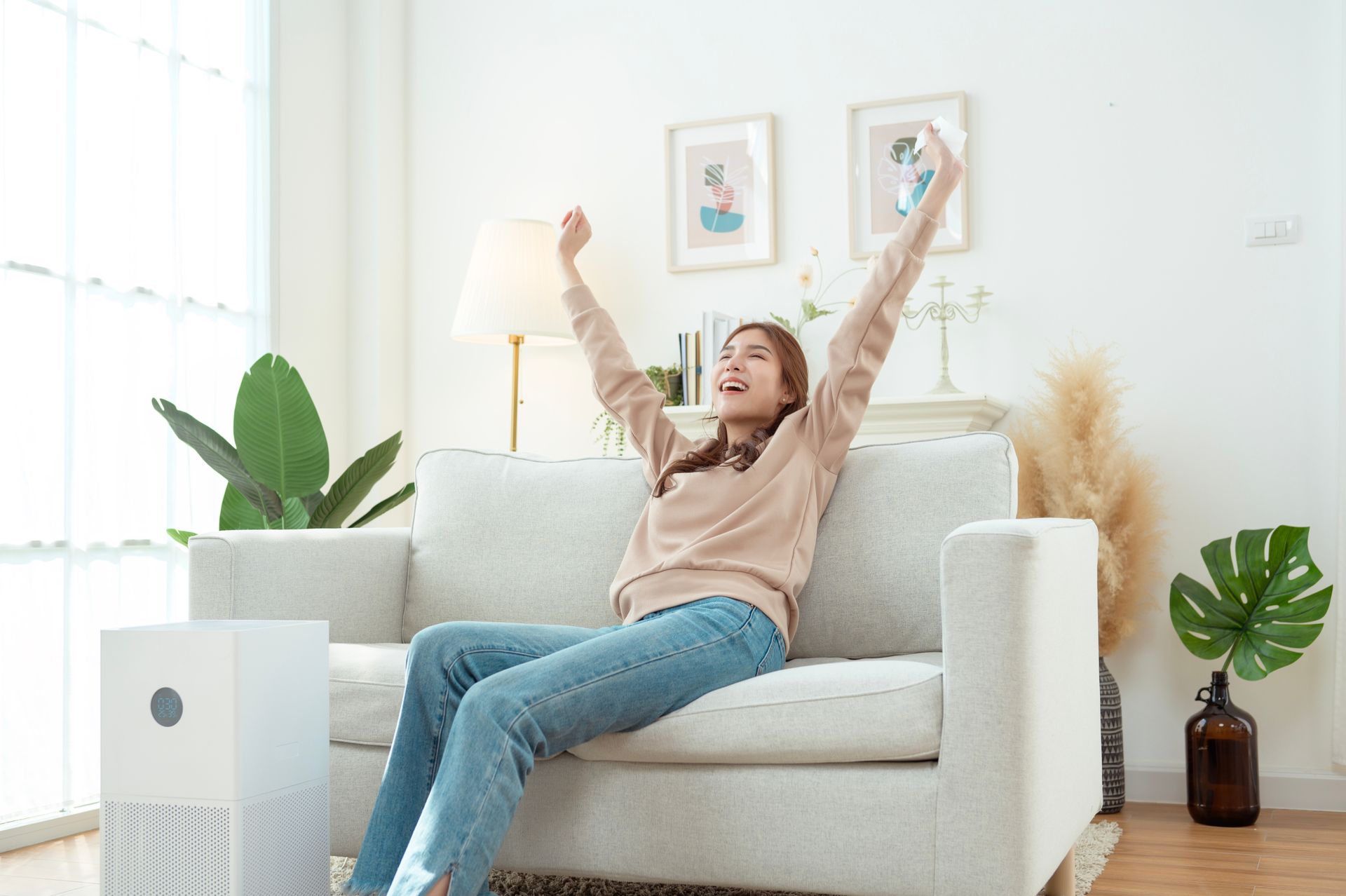 Woman on couch with arms raised, happy expression, near air purifier, plants, and decor.