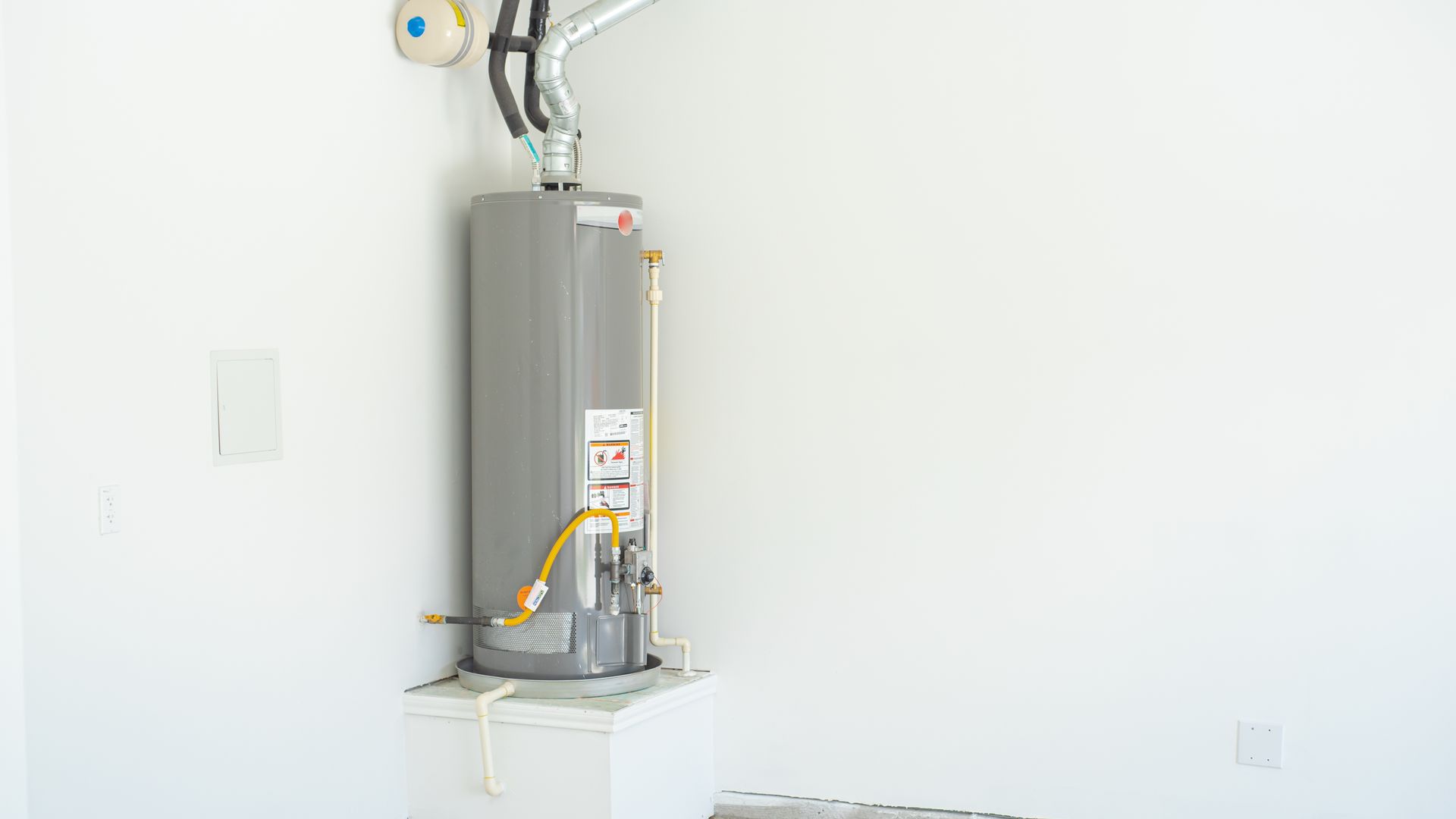Gray water heater against a white wall in an empty room, pipes visible.