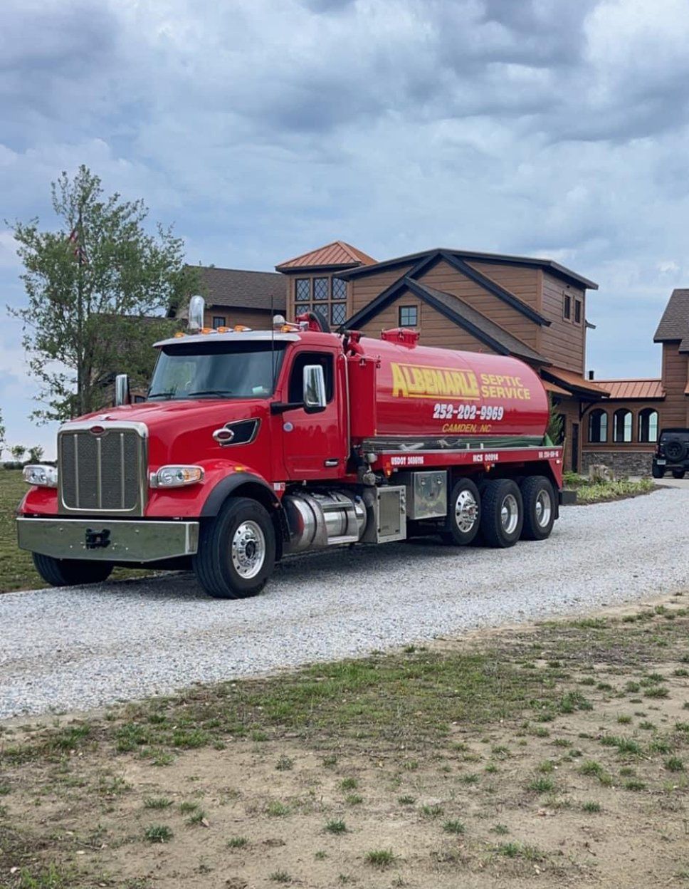 A red tanker truck is parked on a gravel road in front of a house.