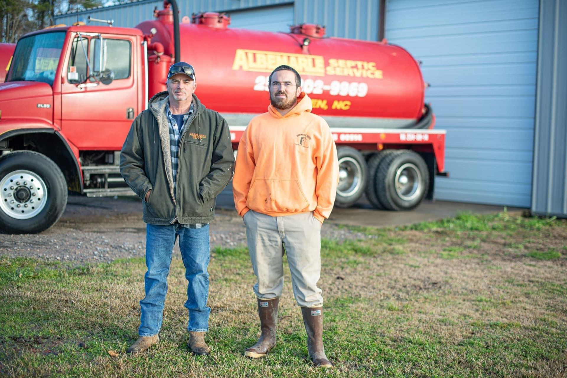Two men are standing in front of a red vacuum truck.