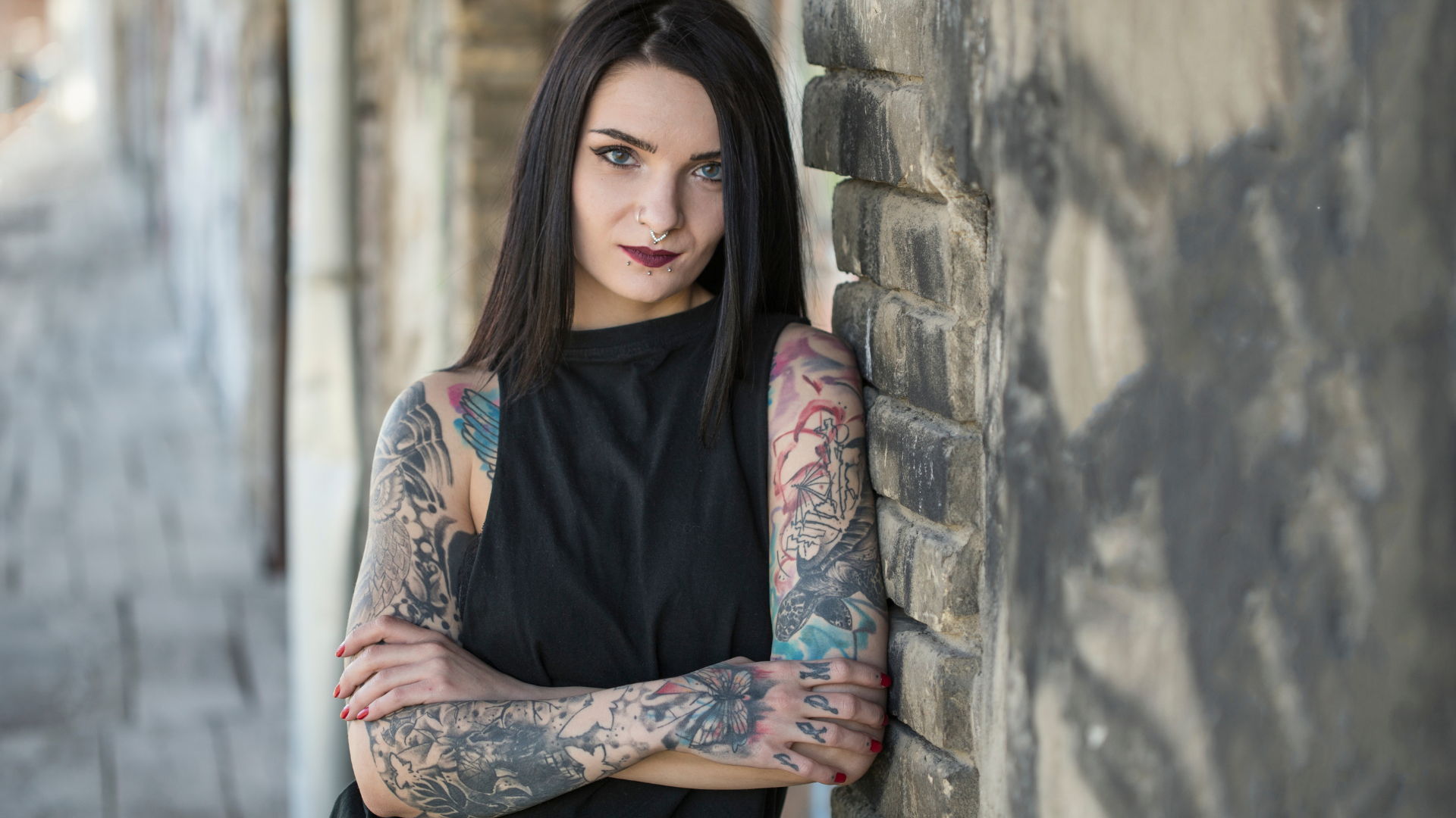 A woman with tattoos on her arms is leaning against a brick wall.
