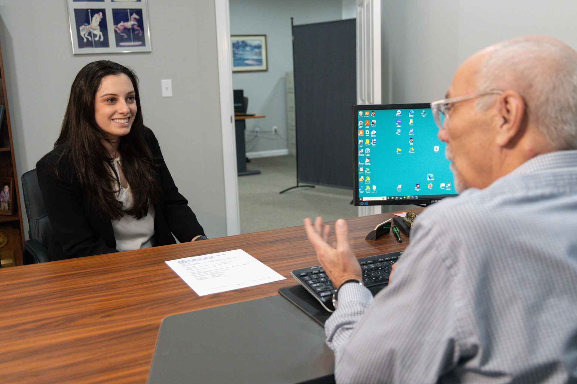 Woman in suit smiles at man; they are in an office. The man gestures toward a computer.