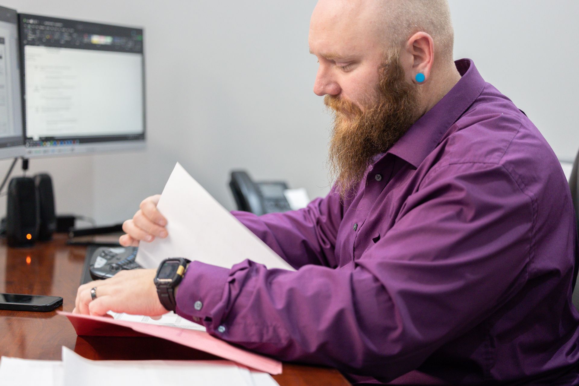 Man with a beard in purple shirt reviews documents at a desk with two computer monitors and a phone.