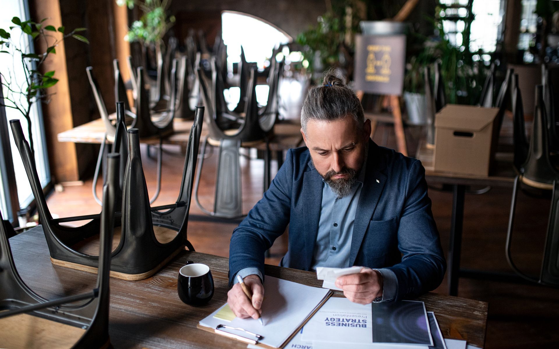 Man in a suit reviews paperwork at a table in an empty restaurant with chairs upturned.