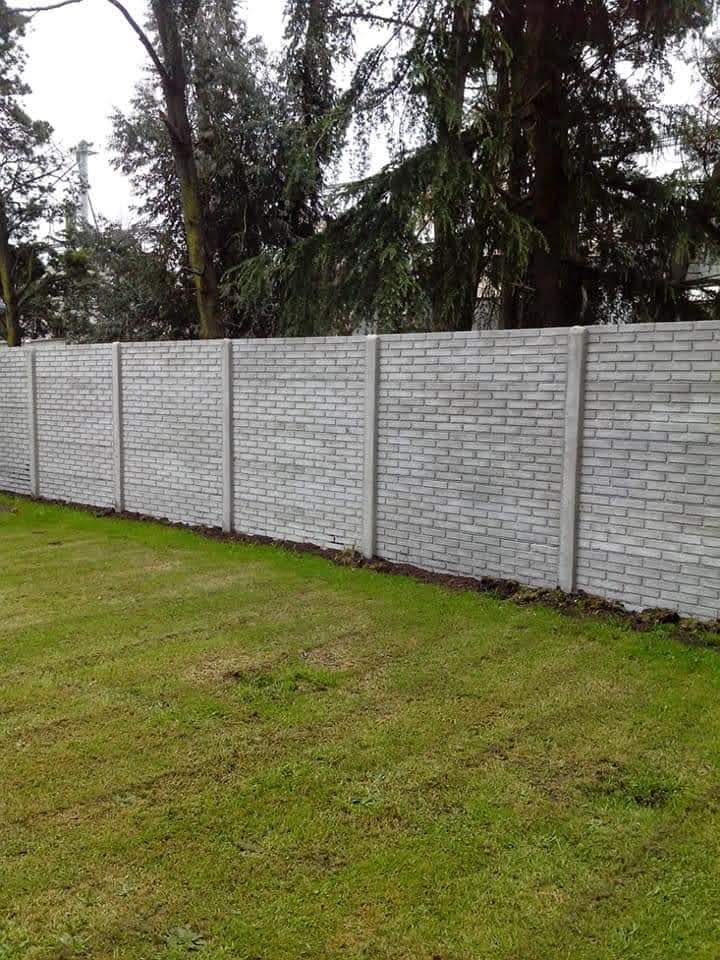 Valla de hormigón gris que bordea un césped verde, árboles al fondo.