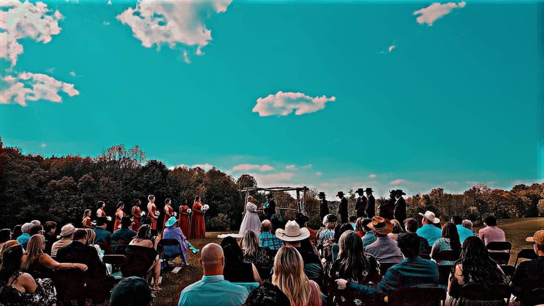 A large group of people are sitting in a field watching a wedding ceremony.