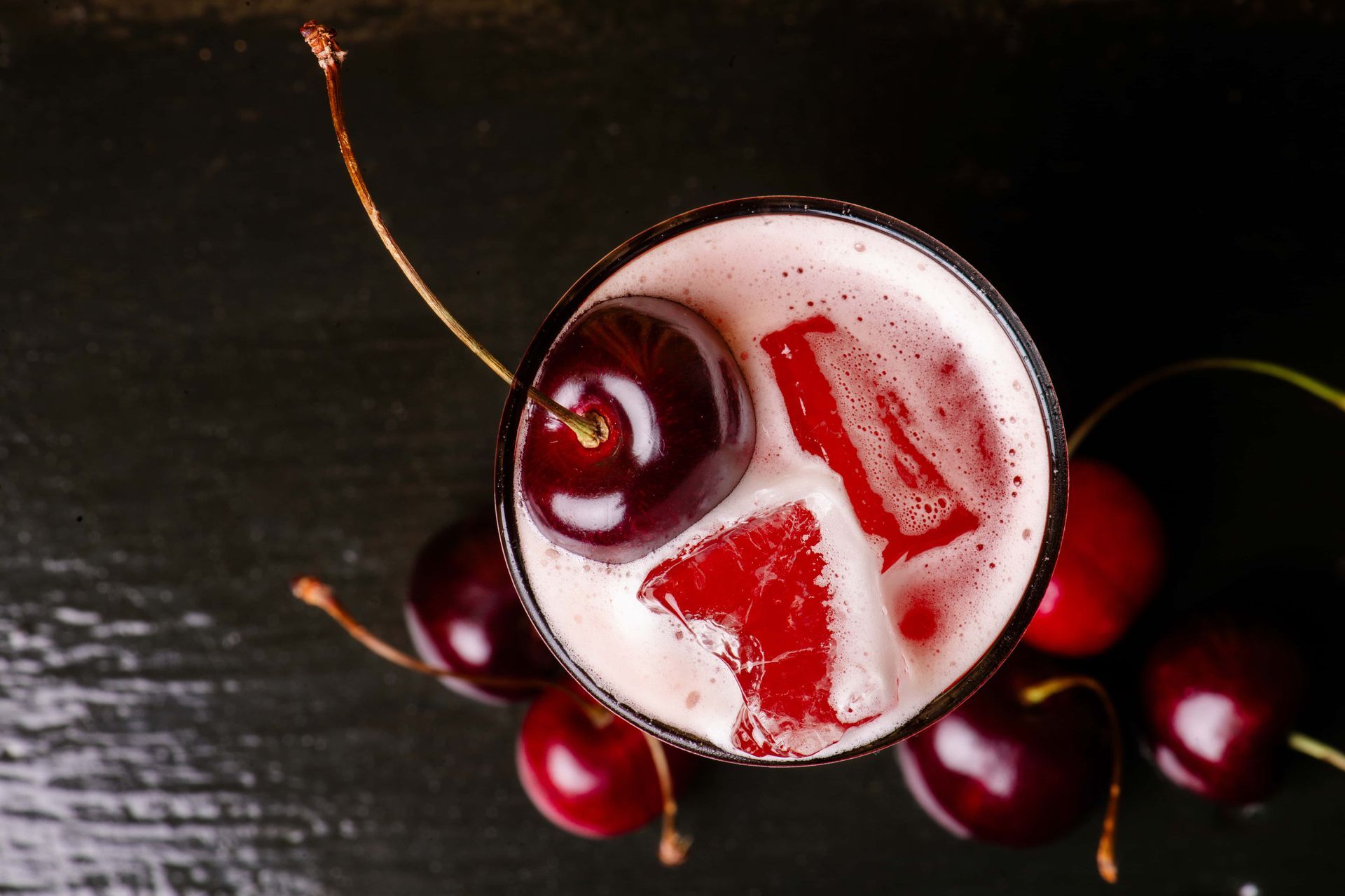 A glass of cherry juice with ice cubes and a cherry on top.