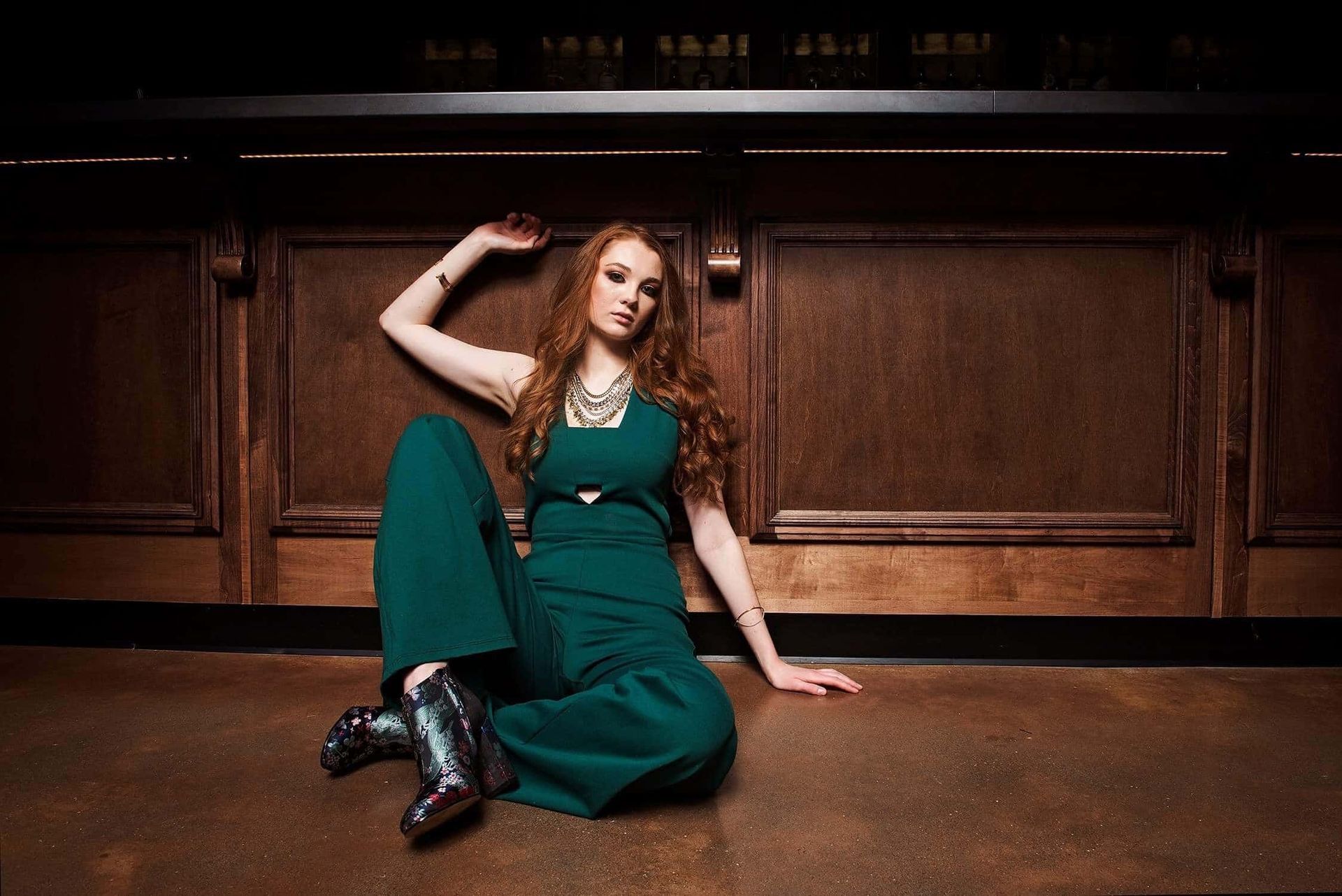 A woman in a green jumpsuit is sitting on the floor in front of a wooden wall.