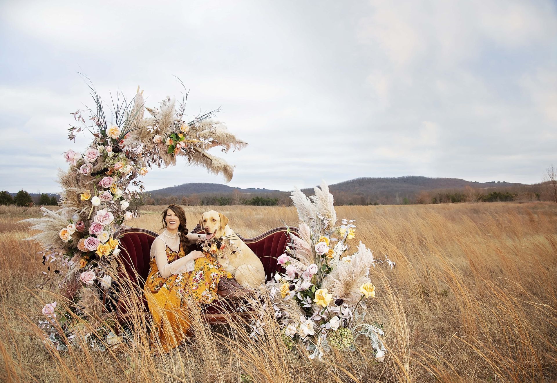 A woman and a dog are sitting on a couch in a field surrounded by flowers.