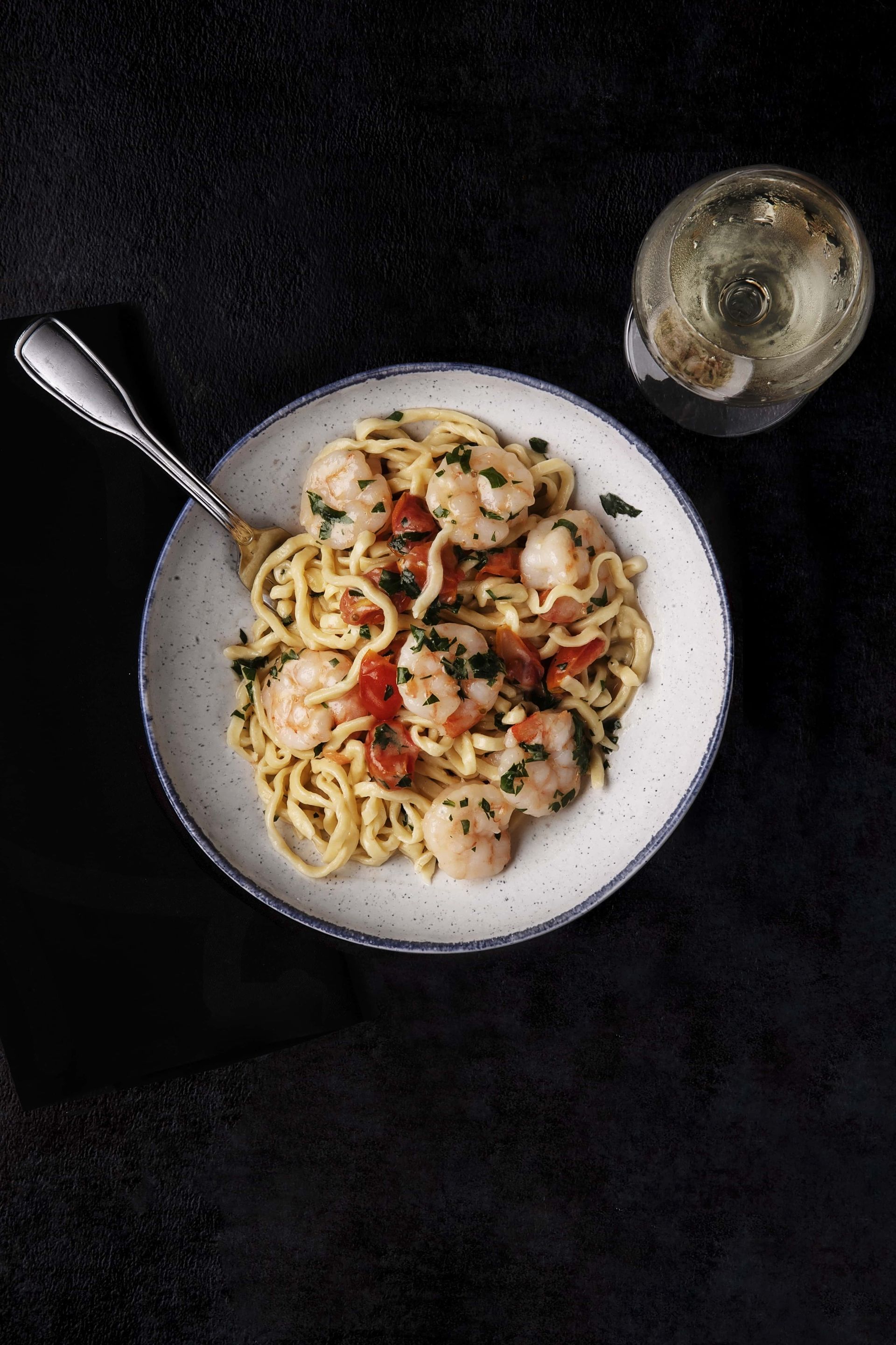 A bowl of pasta with shrimp and tomatoes next to a glass of wine.