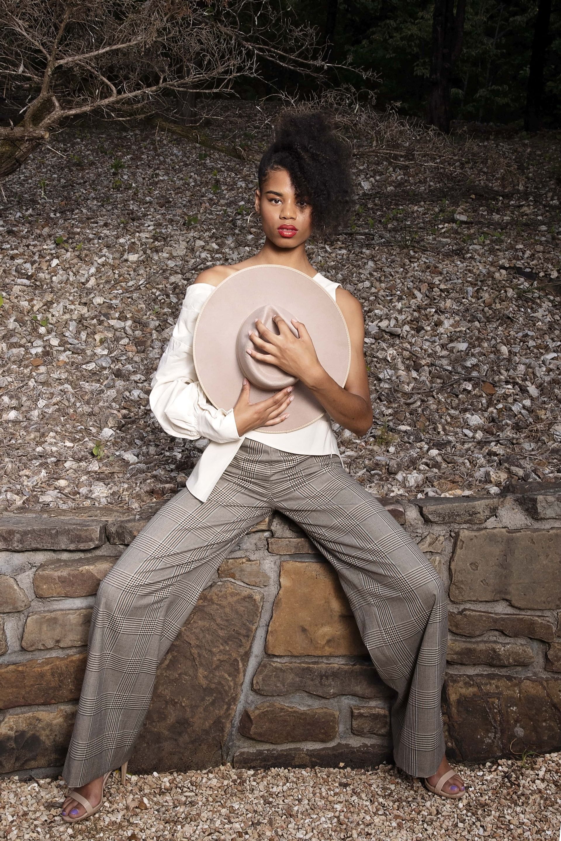 A woman is sitting on a rock wall holding a hat.