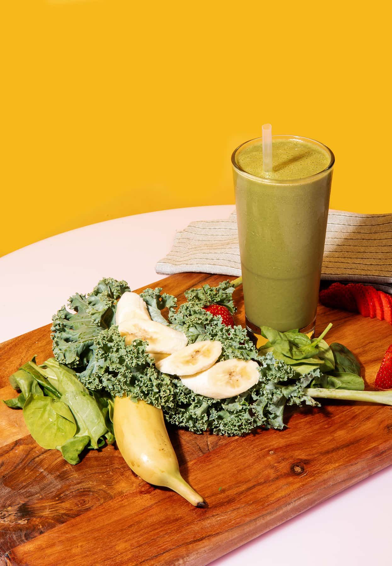 A green smoothie is sitting on a wooden cutting board surrounded by vegetables.