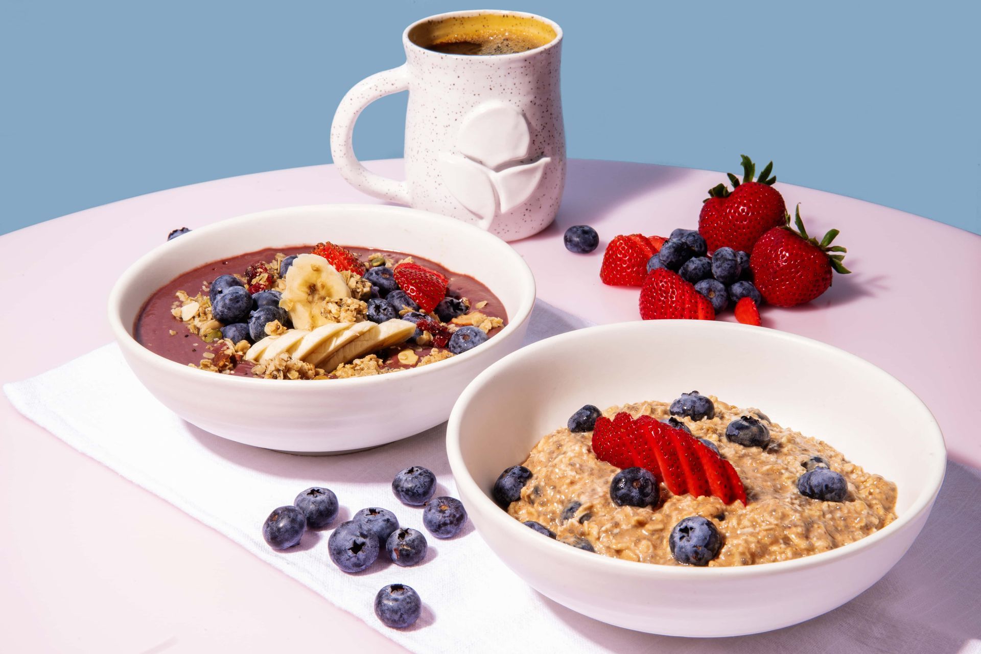 Two bowls of cereal with berries and a cup of coffee on a table.