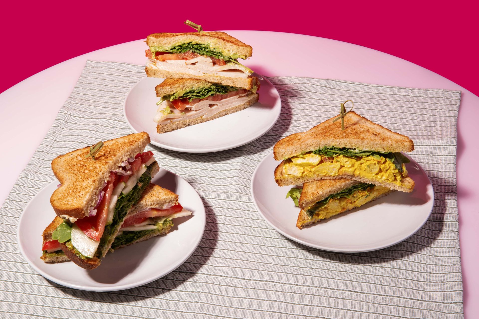 Three sandwiches are on three plates on a table.