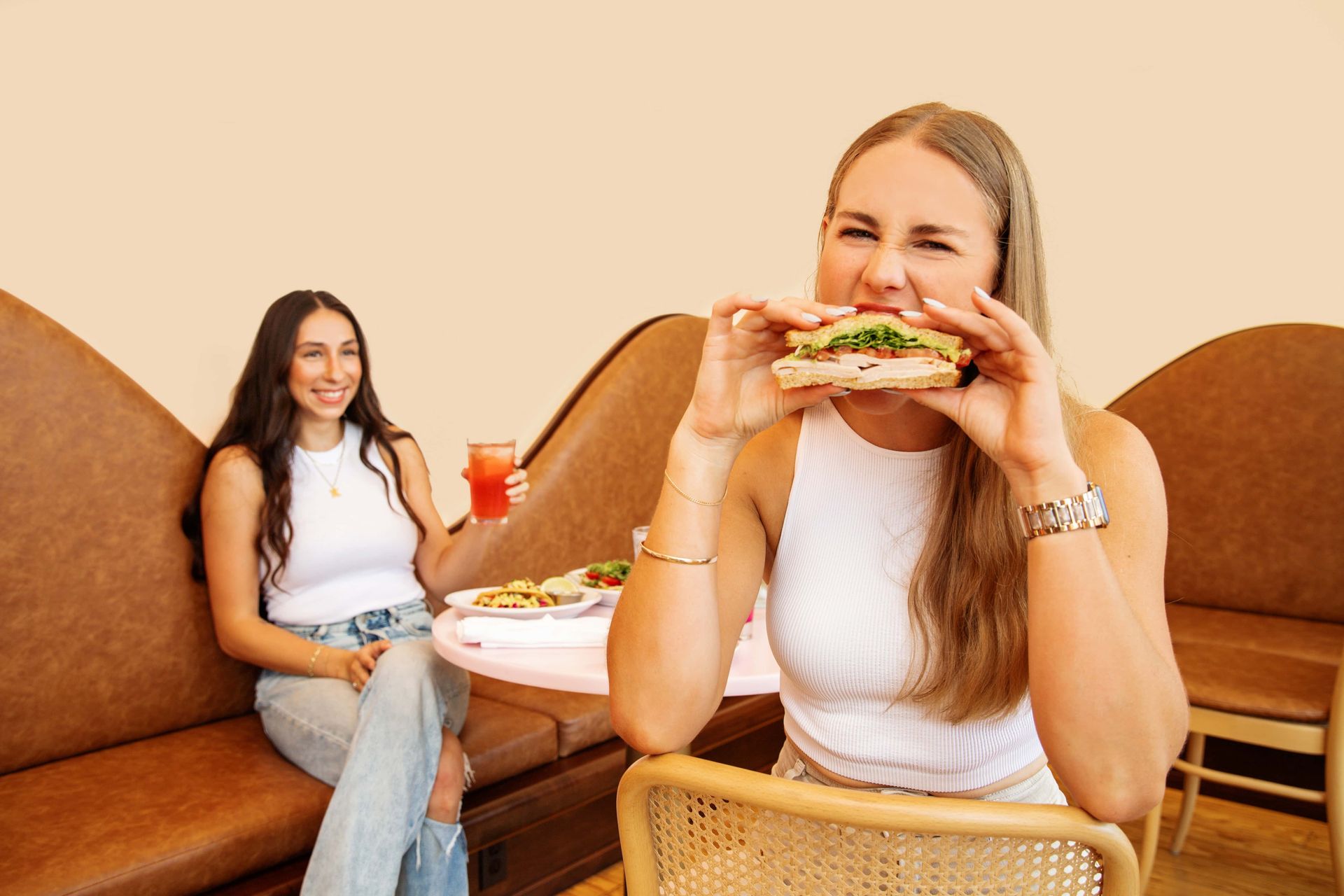 Two women are sitting at a table eating sandwiches and drinking juice.